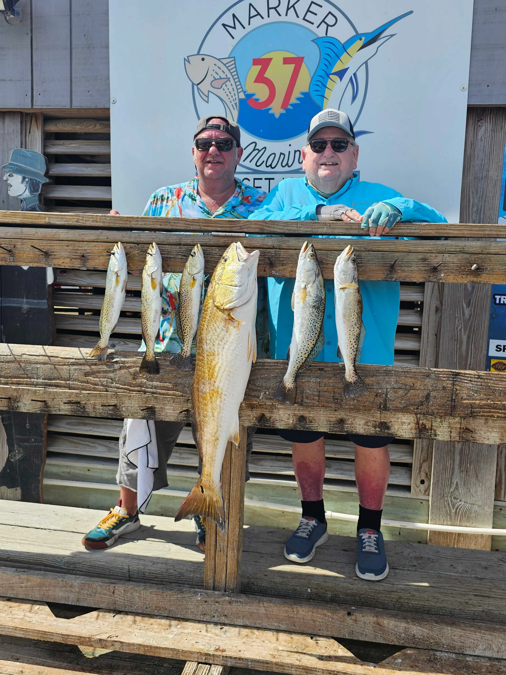 Two men are standing next to a wooden railing holding fish.