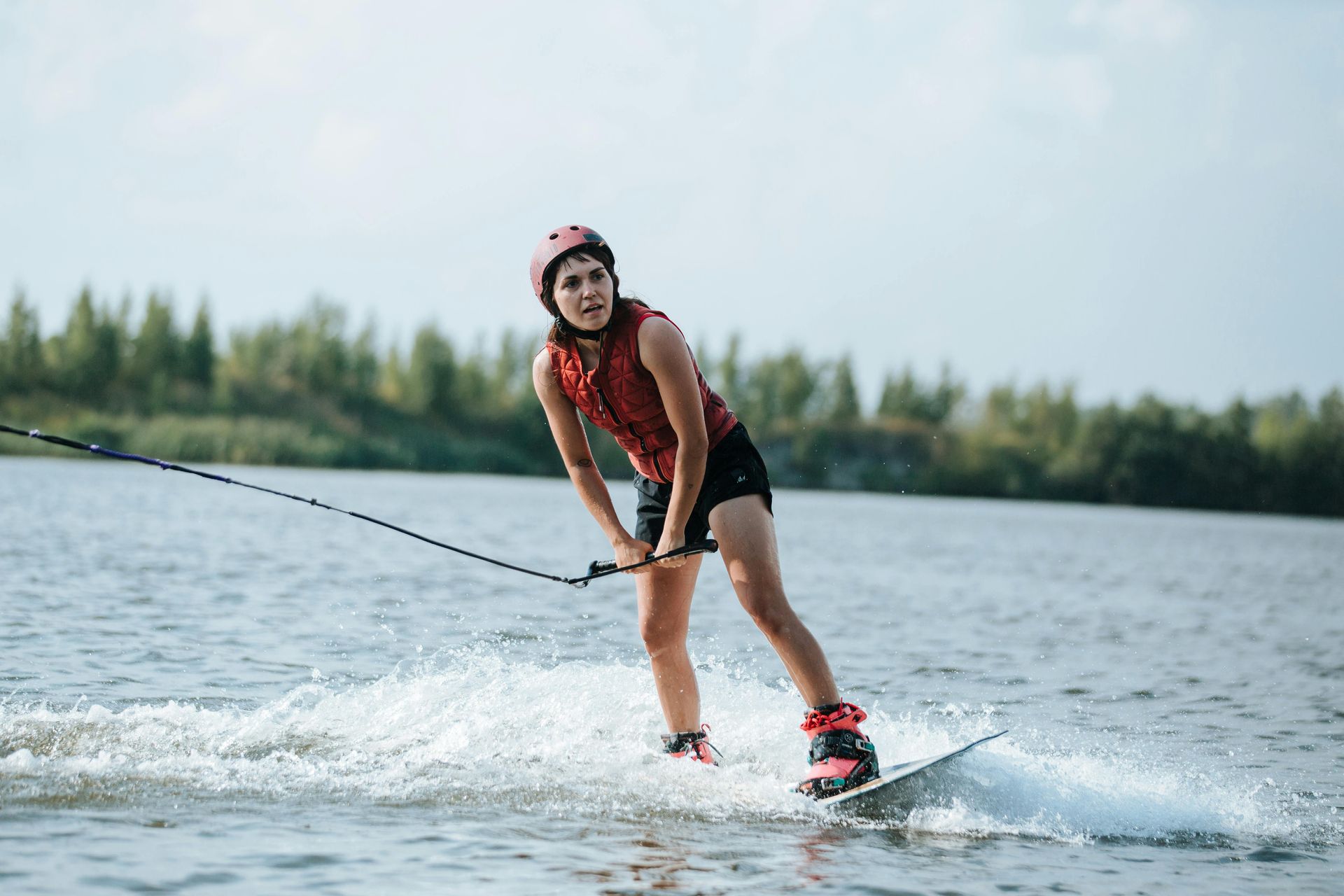 Mujer practicando wakeboard en un lago, sujetando una cuerda. Lleva casco y chaleco salvavidas.
