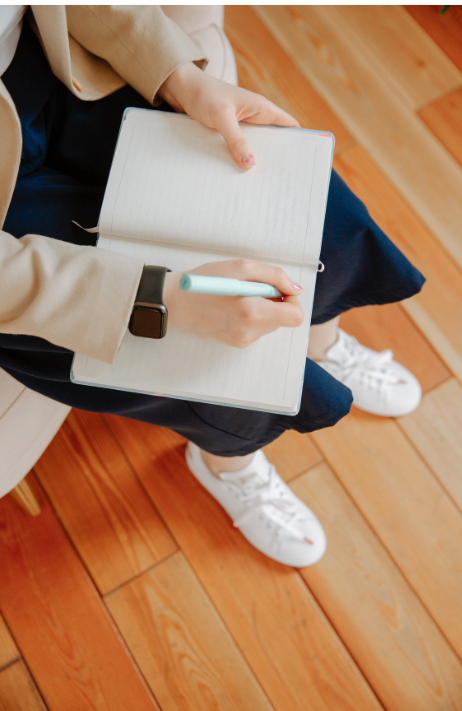 A woman is sitting on a chair holding a notebook and a pen.