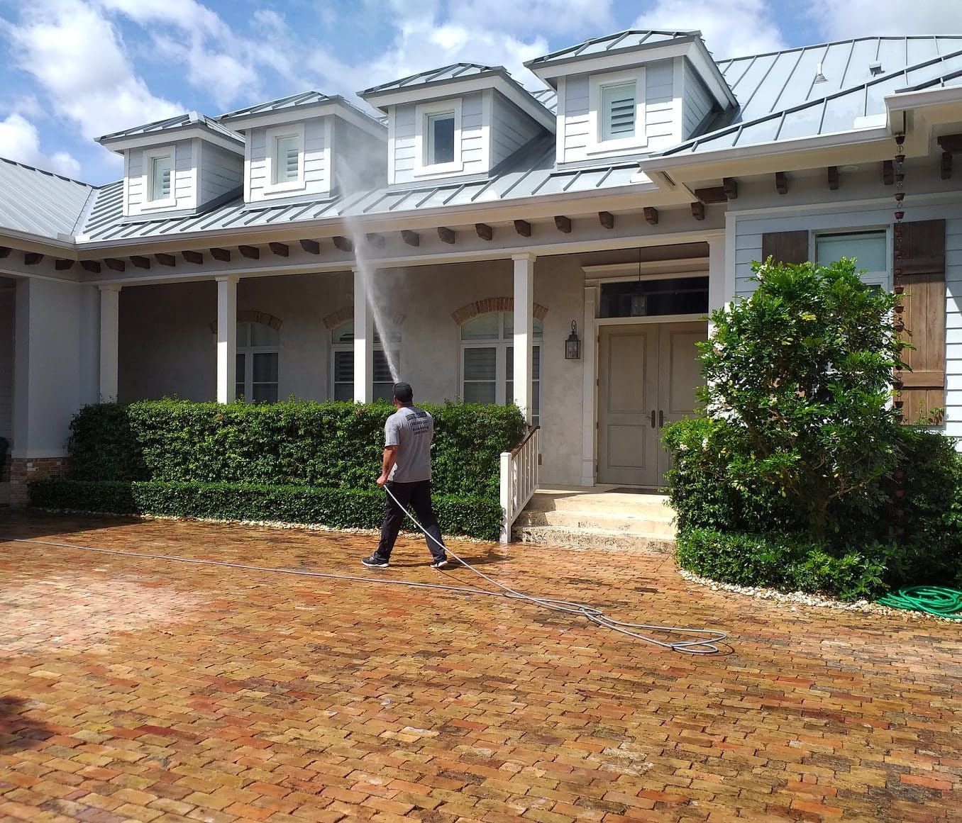 A man is spraying water on a brick driveway in front of a large house.