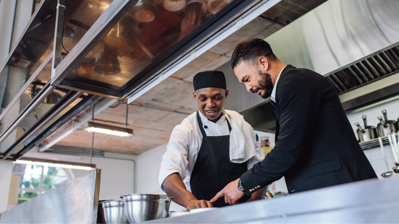 A chef and a person in a suit discuss work in a professional kitchen, with the person in the suit pointing at a surface.
