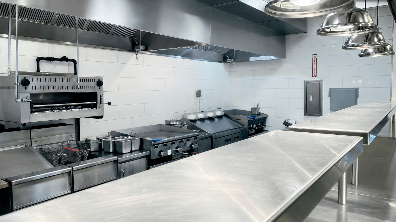 Empty commercial kitchen with stainless steel counters, sinks, stovetops, and hanging lights