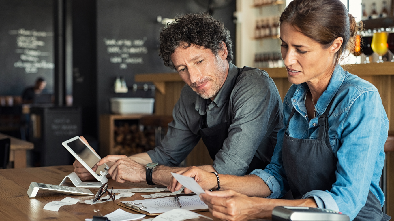 Two coworkers review papers and a tablet at a café table.