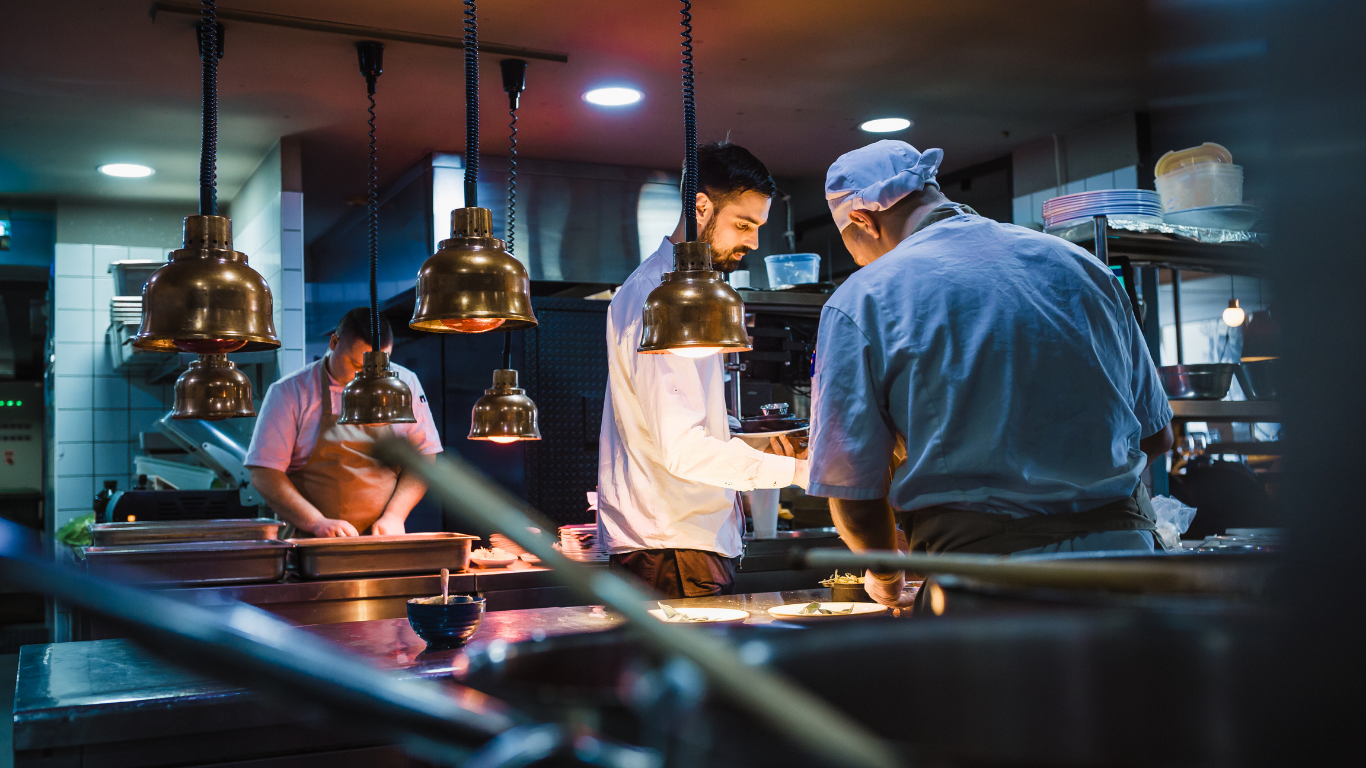 Professional chefs in a dimly lit, industrial kitchen work at prep stations under brass hanging pendant lights.
