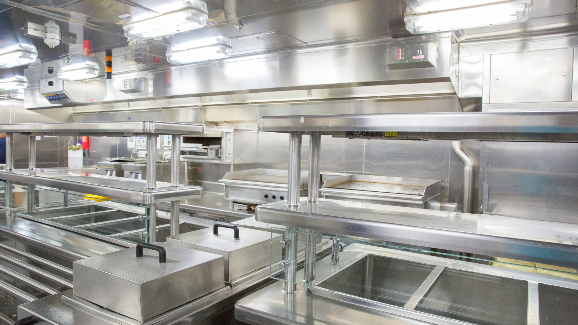 Stainless steel commercial kitchen with vent hoods, prep areas, and bright lights.
