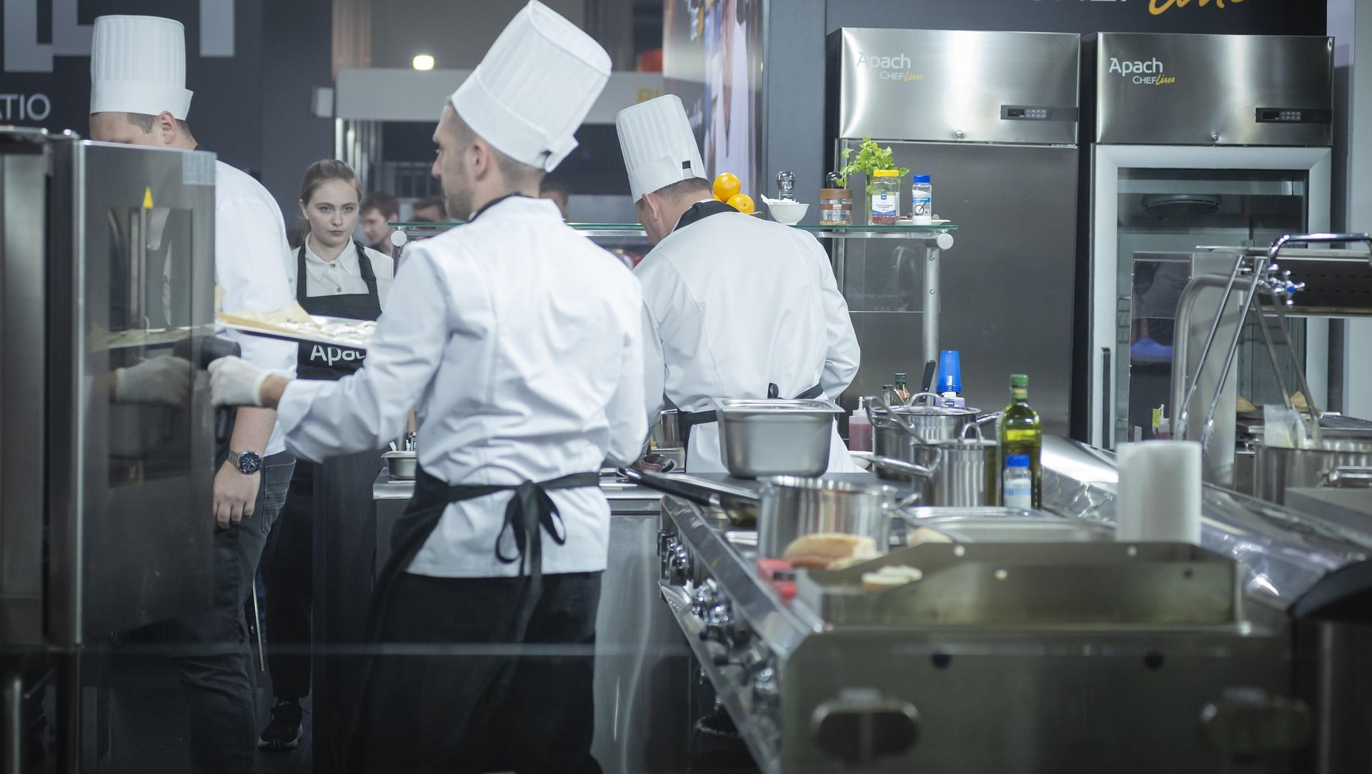 Chefs in white uniforms and hats cooking in a commercial kitchen with stainless steel appliances.