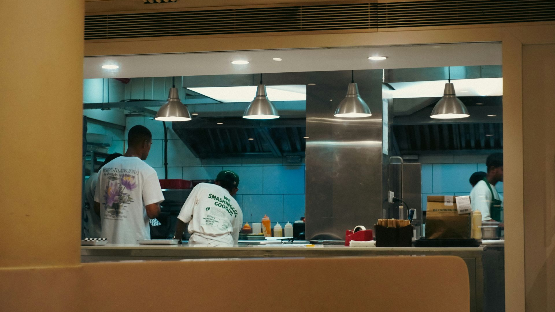 Restaurant kitchen staff in uniforms work behind a service counter under hanging pendant lights.