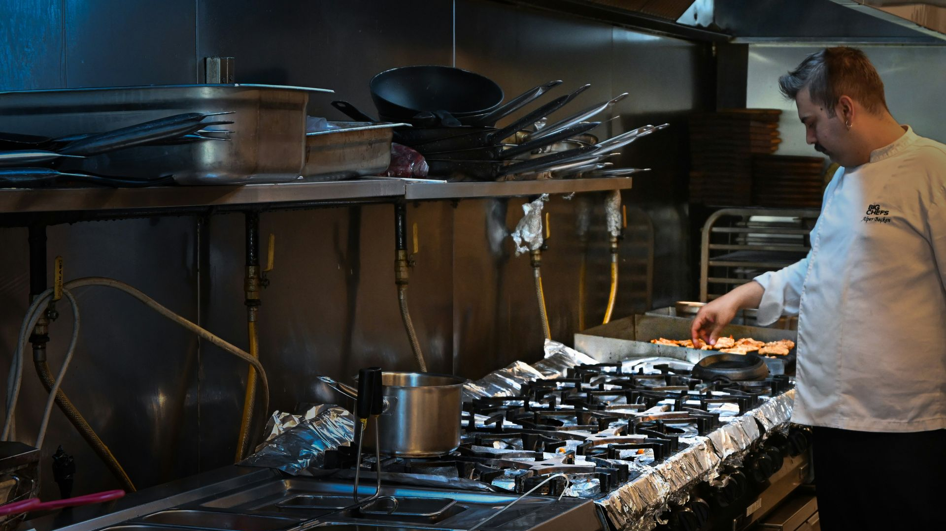A professional chef in a white uniform prepares food on a stove in a commercial kitchen with stainless steel surfaces.
