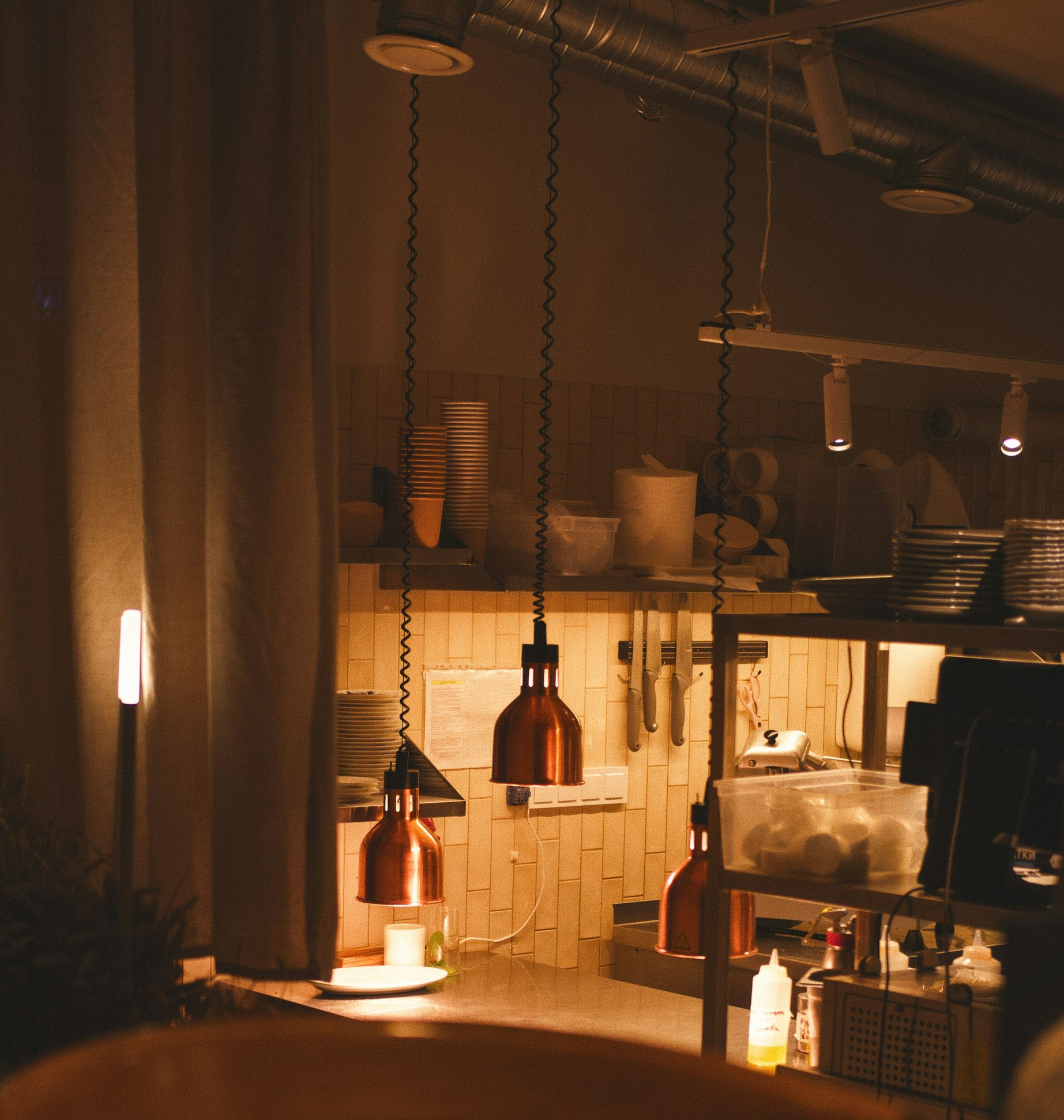 Cozy interior scene with hanging copper pendant lights, wooden table and chair, and a view of a kitchen area.