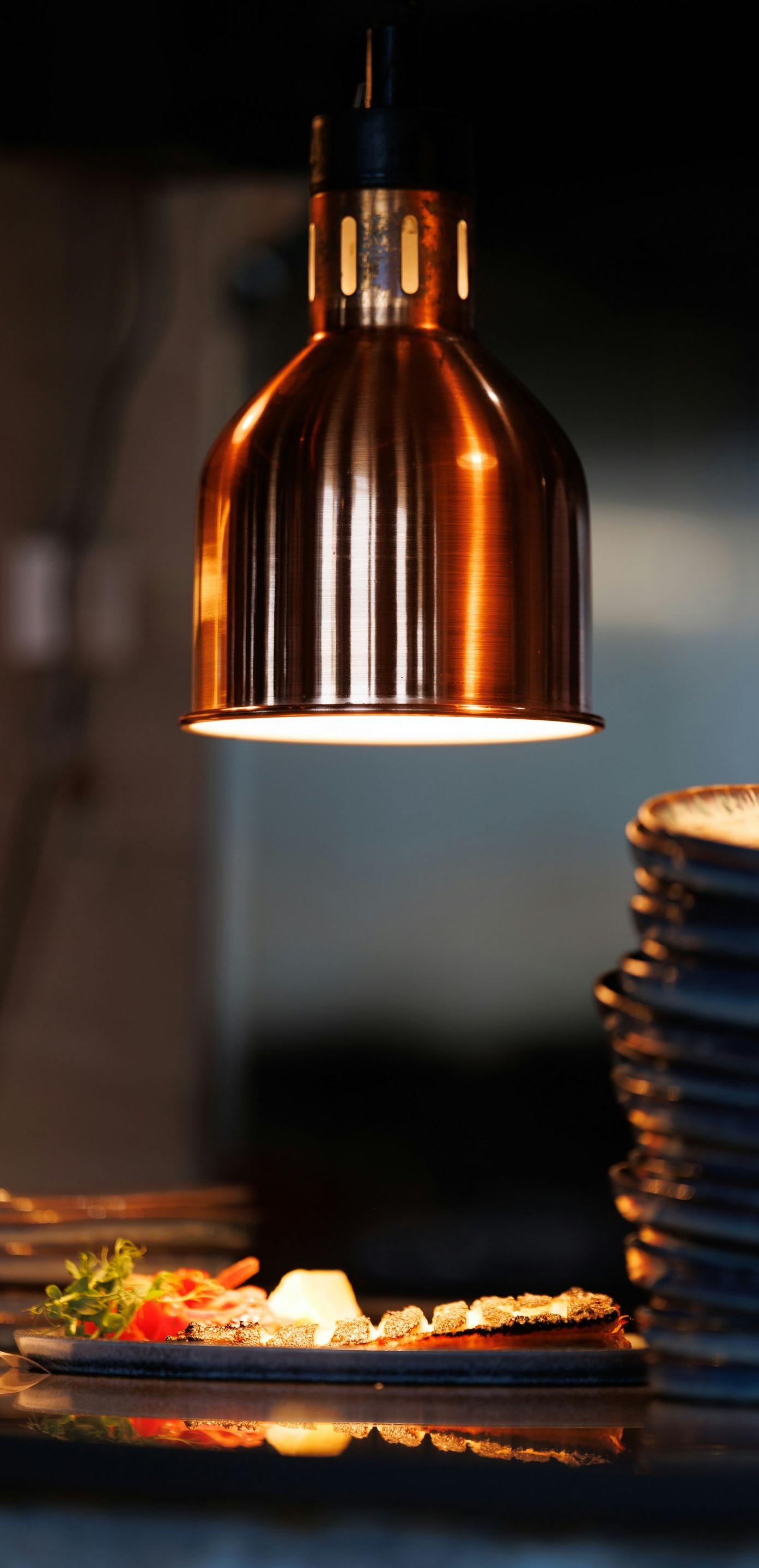 A copper heat lamp hangs above a plated dish and a stack of ceramic bowls in a dimly lit restaurant kitchen.
