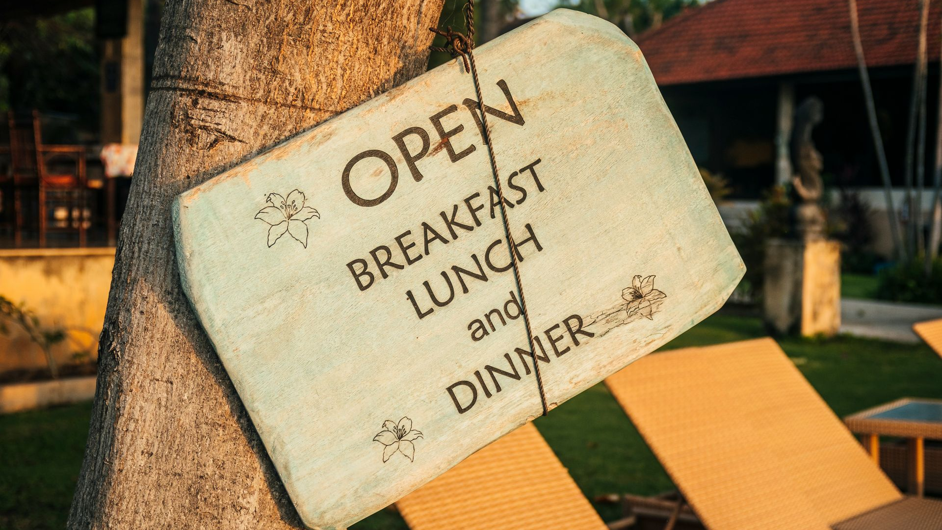 Open sign for breakfast, lunch, and dinner hanging on a tree outdoors near lounge chairs