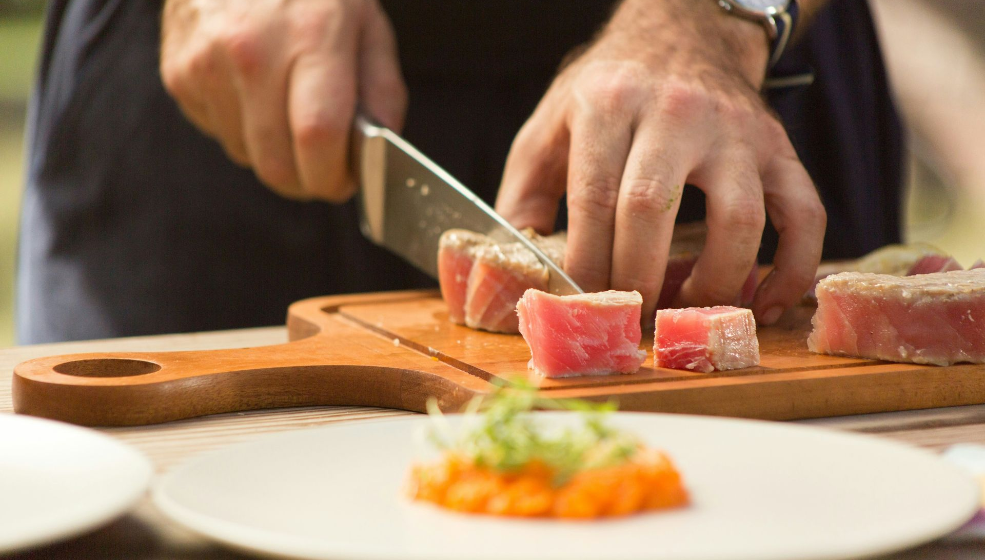 Person slicing raw tuna on a wooden board; plated dish of food in foreground.