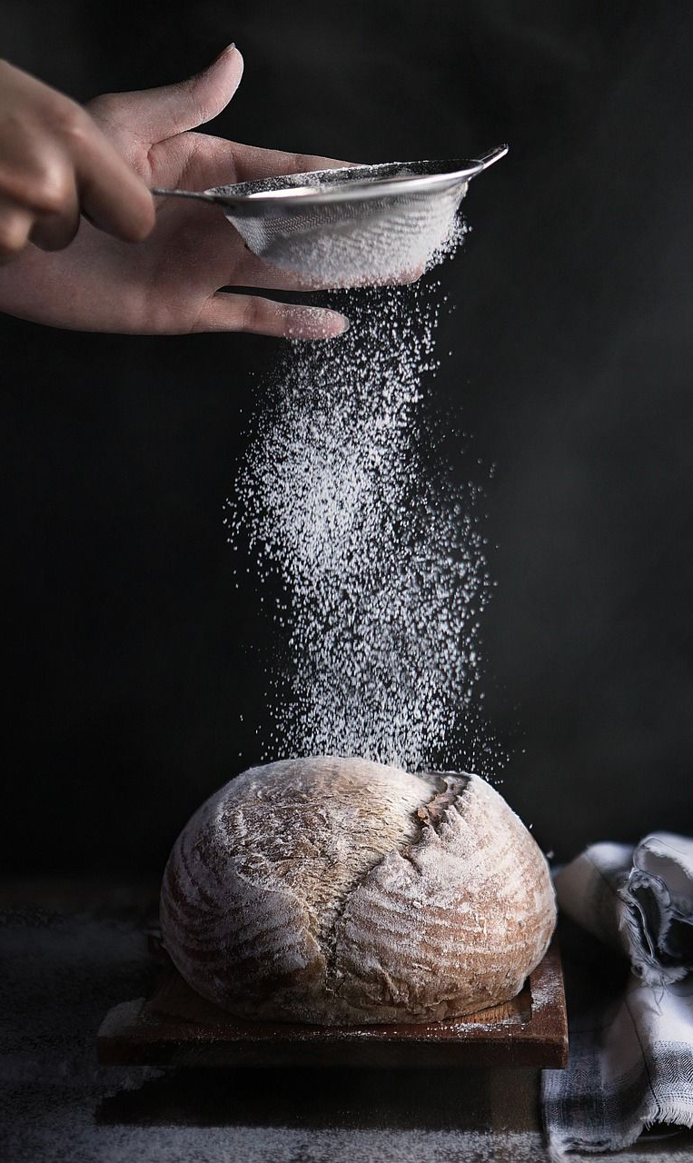 Hand dusting powdered sugar over a loaf of bread with a sifter. Dark background.