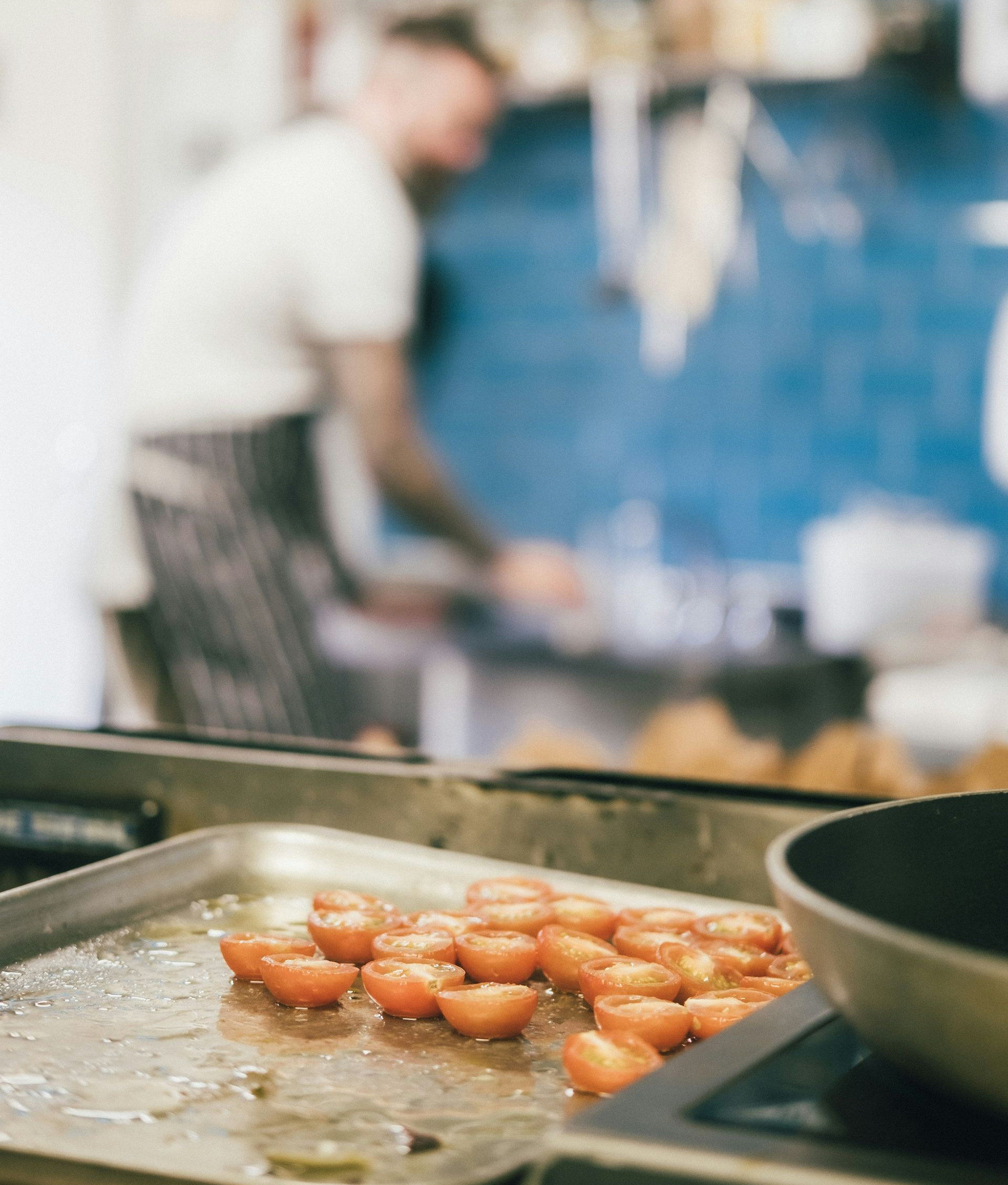 A metal tray of halved, roasting cherry tomatoes sits in the foreground with a blurred chef working in a kitchen behind.