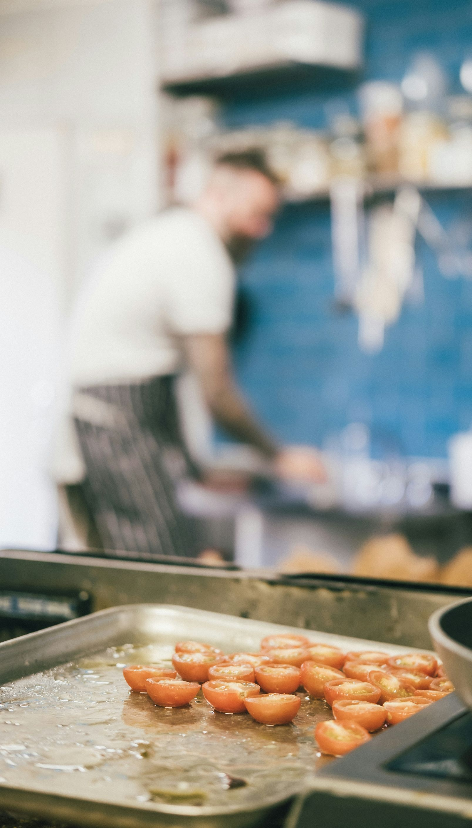 Halved tomatoes on a baking sheet in a kitchen, chef blurred in background.