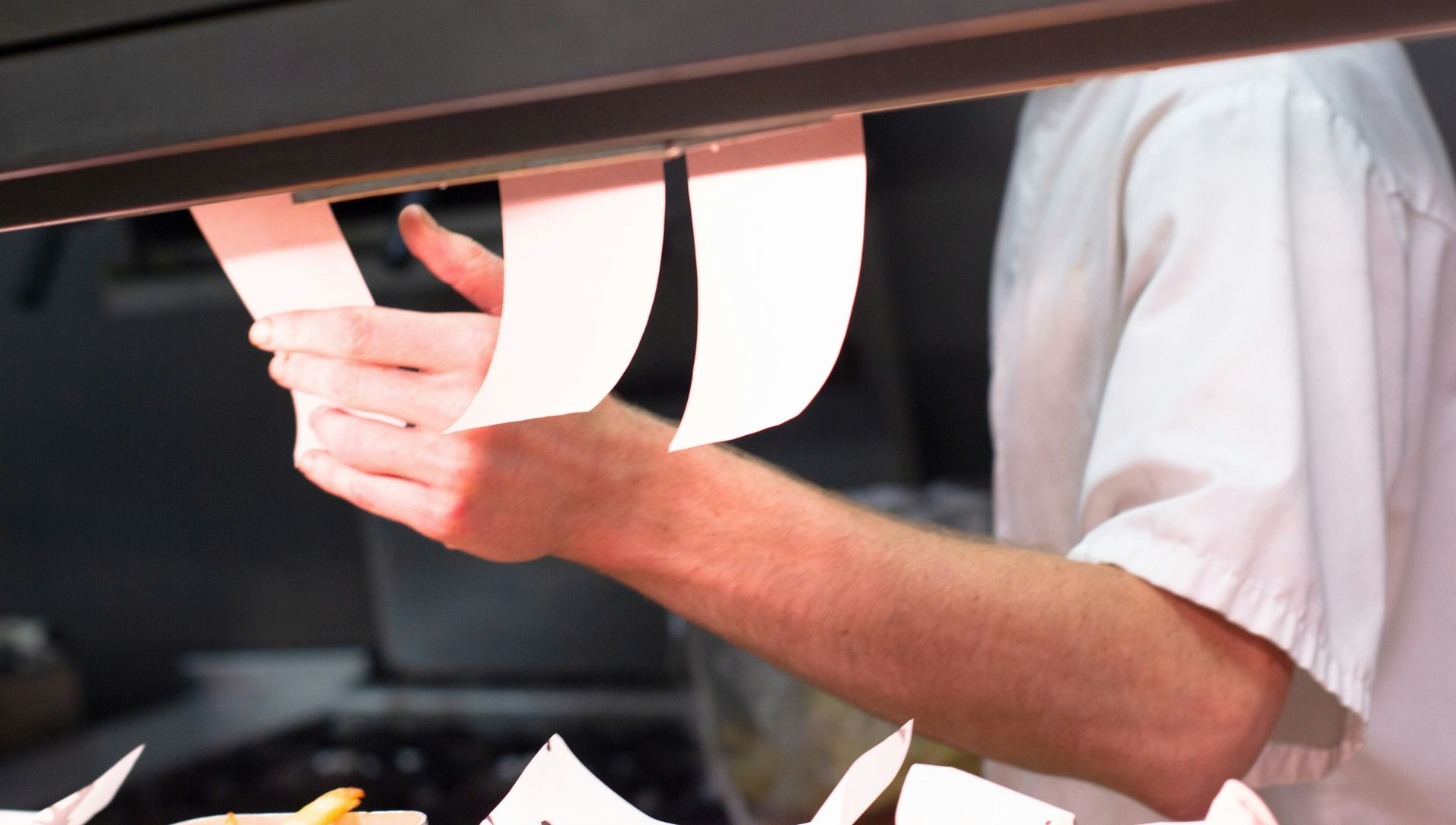 Person in white shirt placing order tickets on a restaurant kitchen rail.