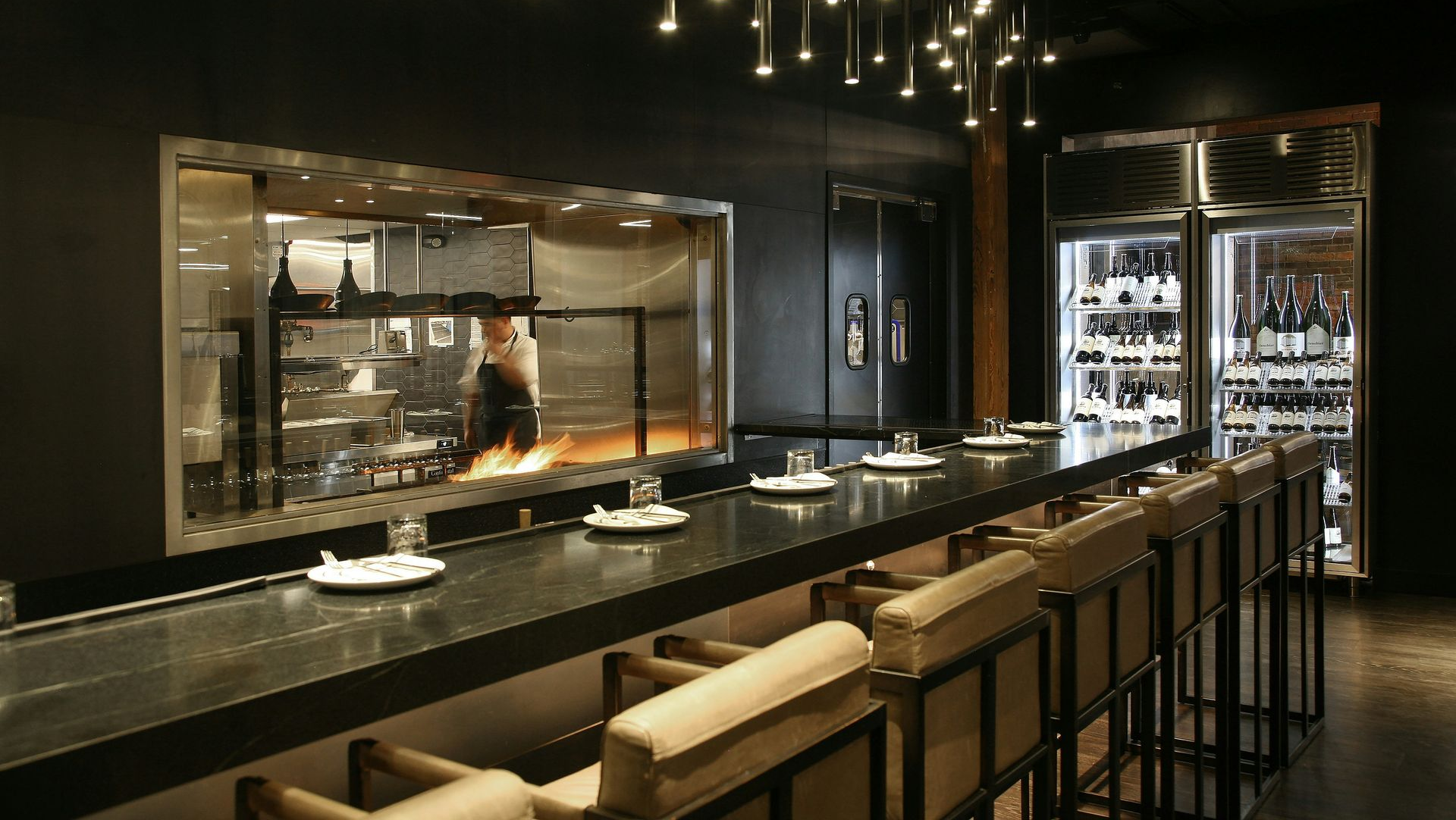 Restaurant interior with a dark bar, open kitchen with flames, and hanging lights.
