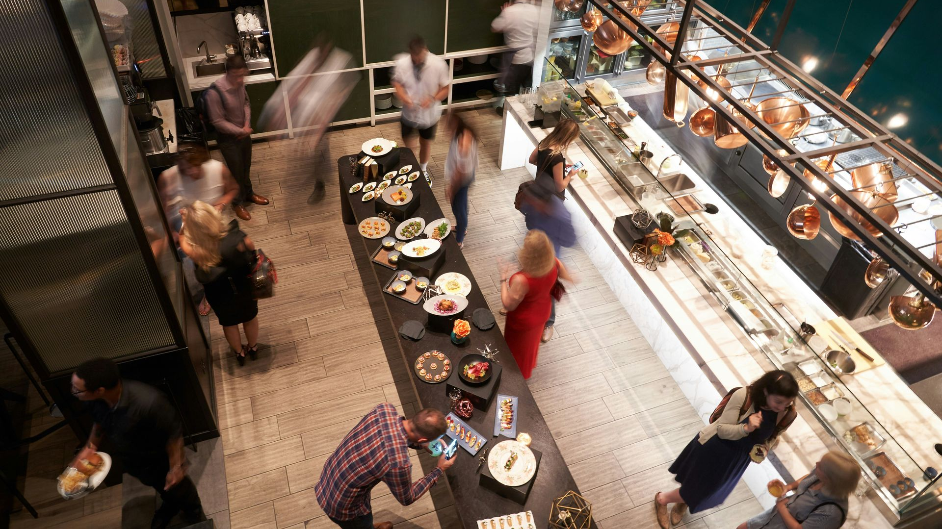 Event at a restaurant: people mingling near a long food table, bar above, copper lights.