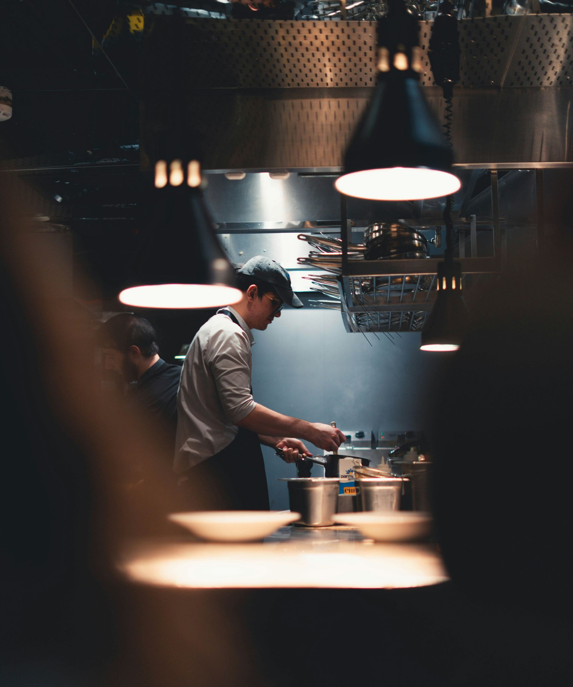 Chef cooking in a dimly lit kitchen under hanging lights, viewed through a blurred foreground.