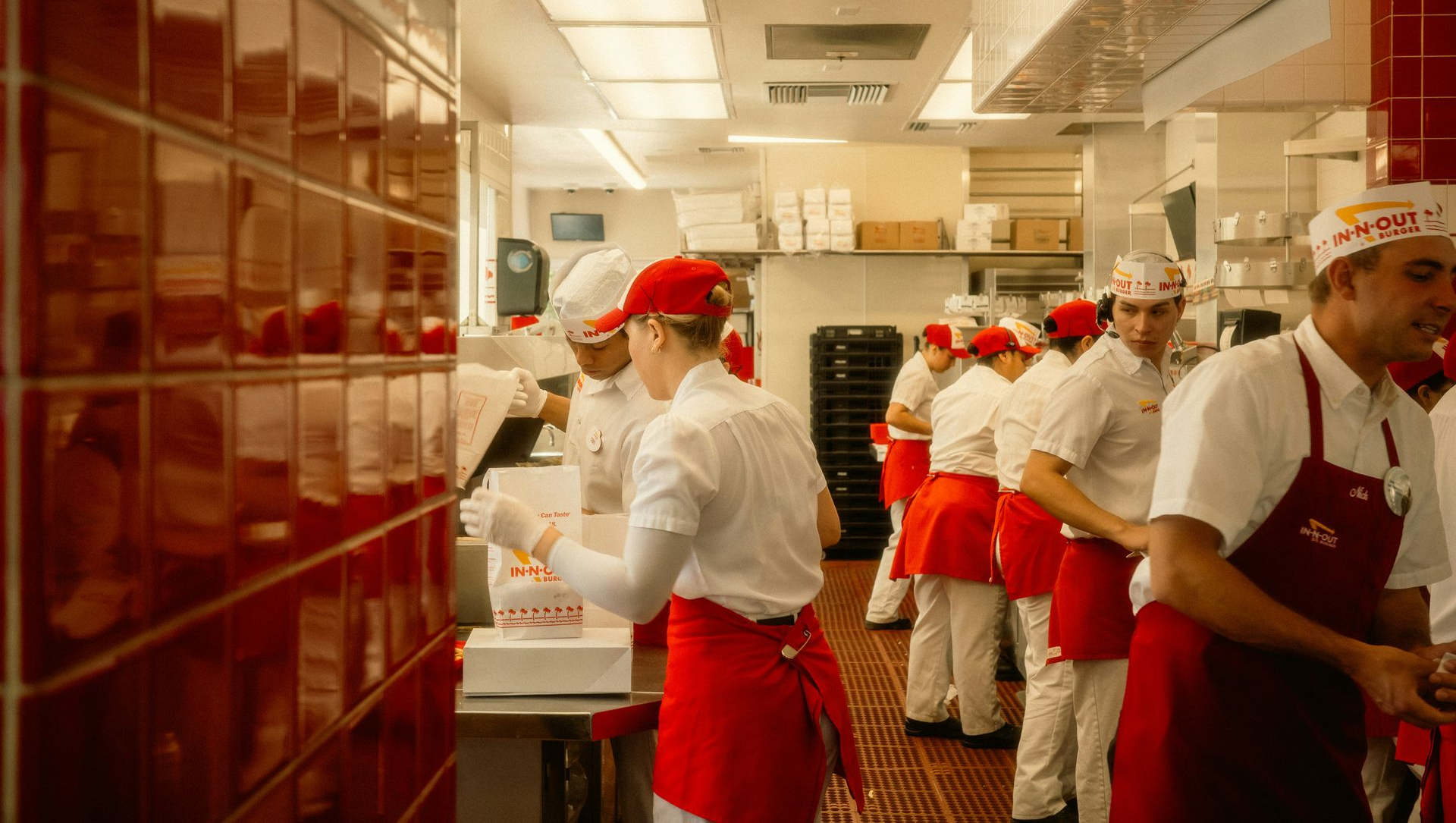 Chefs in red aprons work in a bright commercial kitchen with white tiled walls and counters