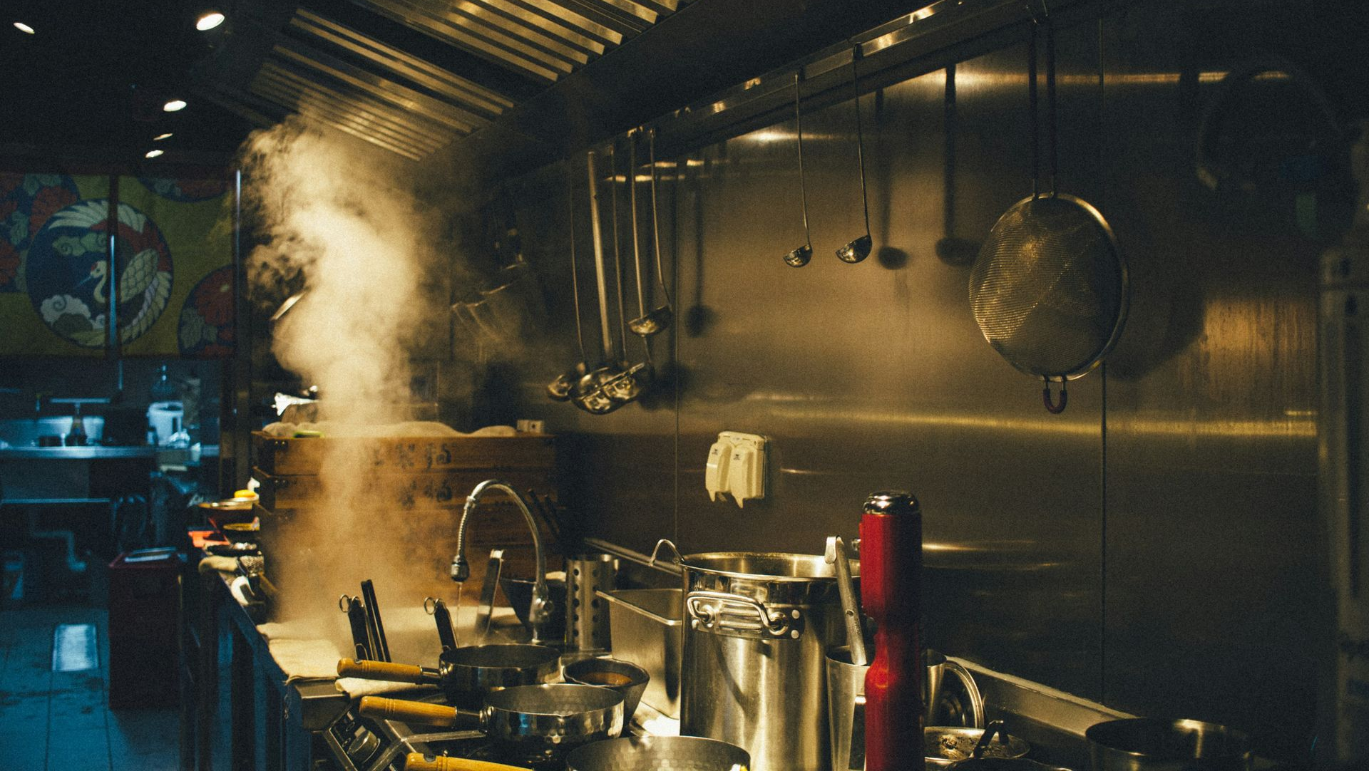 Steaming restaurant kitchen with stainless steel appliances and utensils, smoke rising.