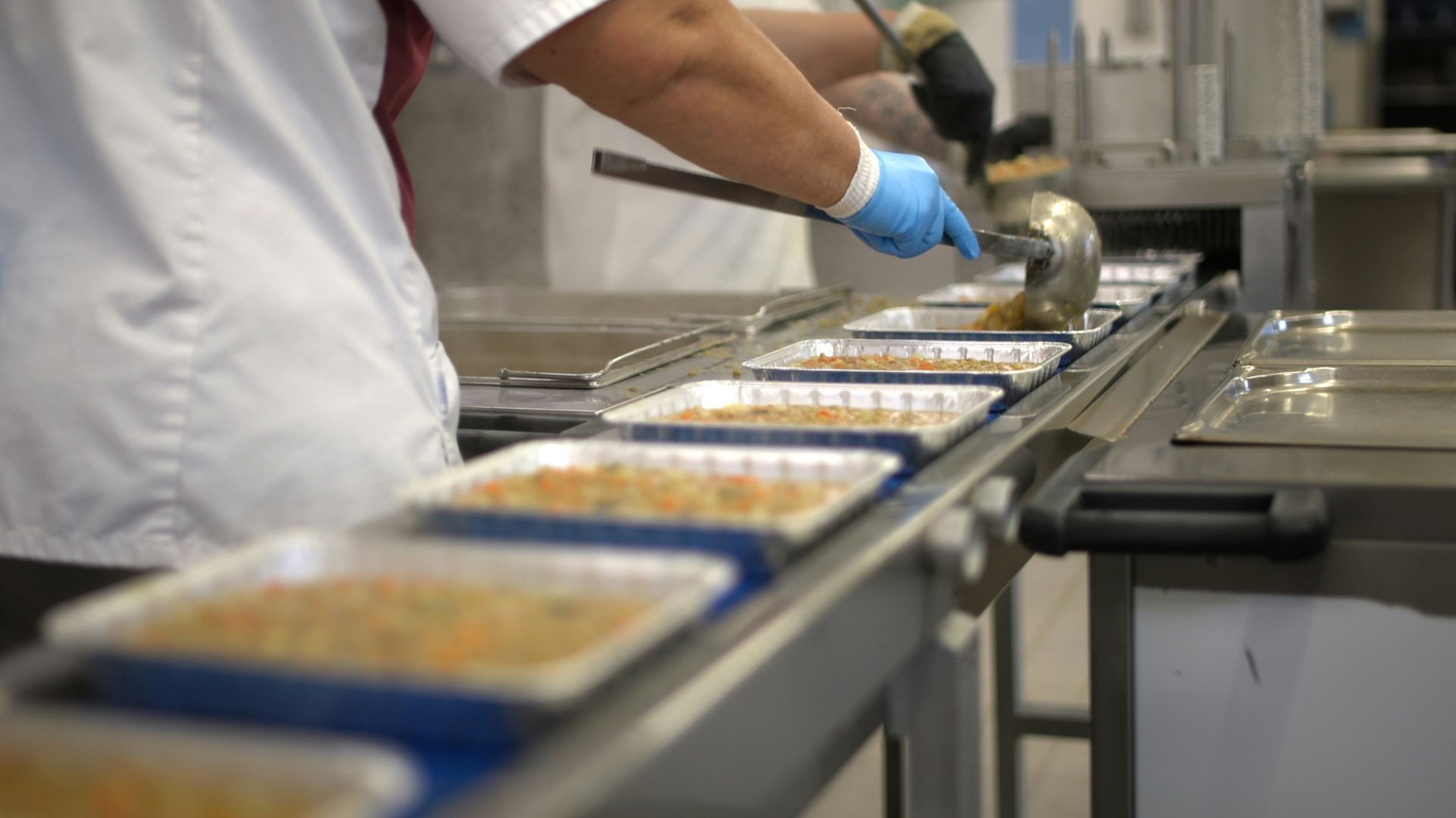 A person wearing a white uniform and blue gloves uses a ladle to portion food into trays on a moving assembly line.