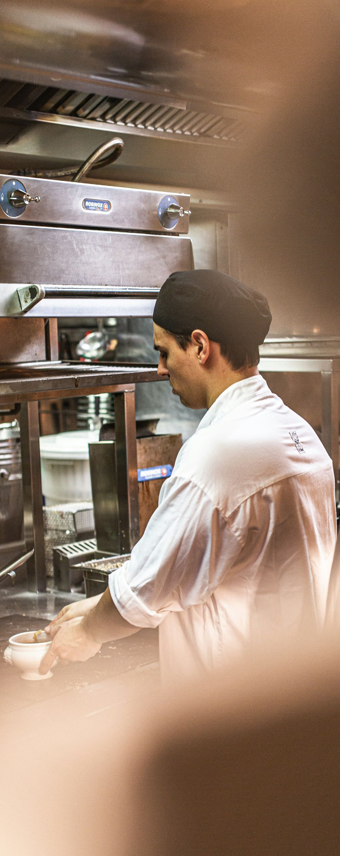 Chef in white coat working at a stainless-steel kitchen station with steam rising around him.