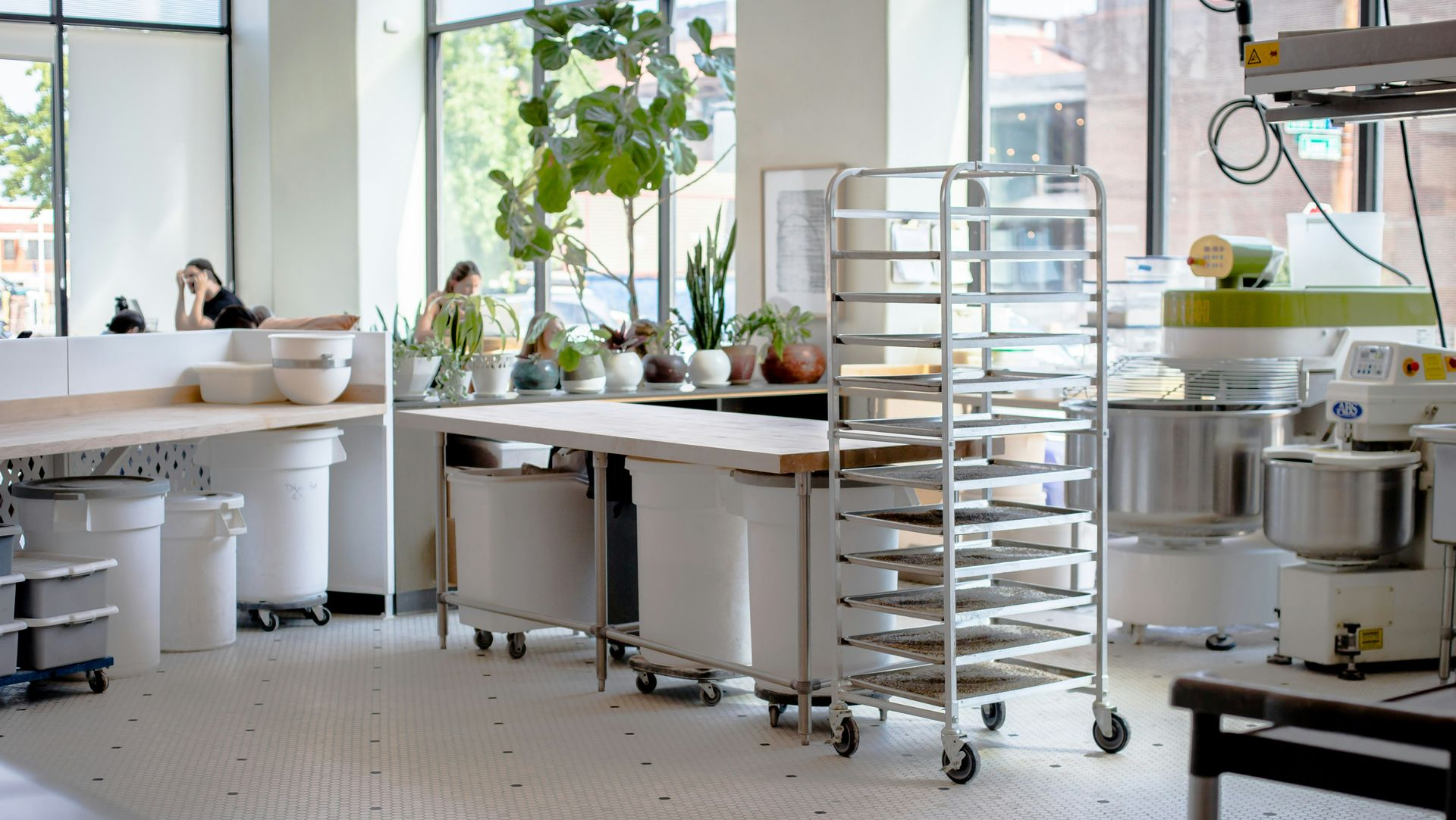 A bright, airy commercial kitchen features metal racks, industrial mixers, and work tables on a light, patterned floor.