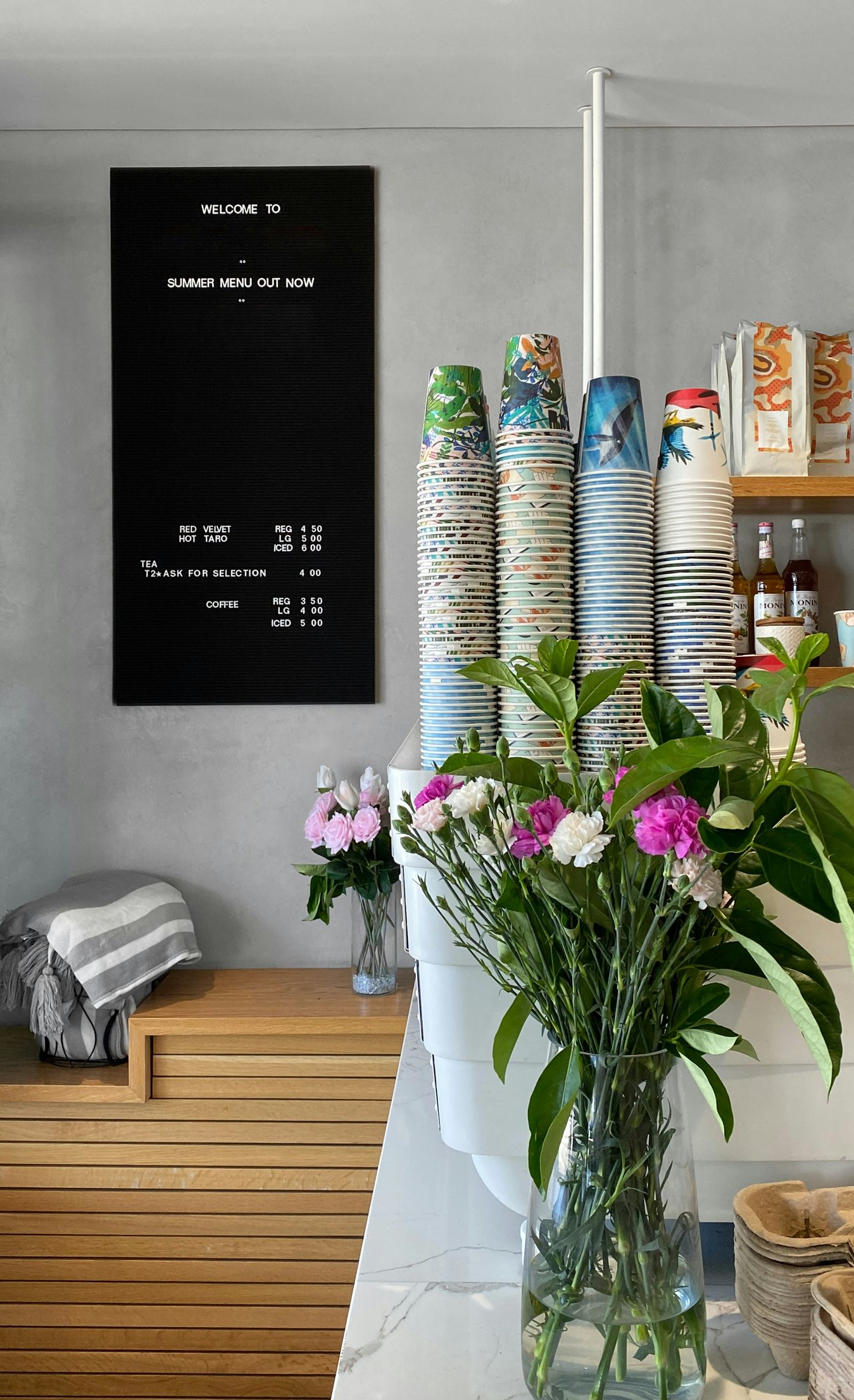 Cafe interior with menu board, stacked cups, flowers, and wooden counter.