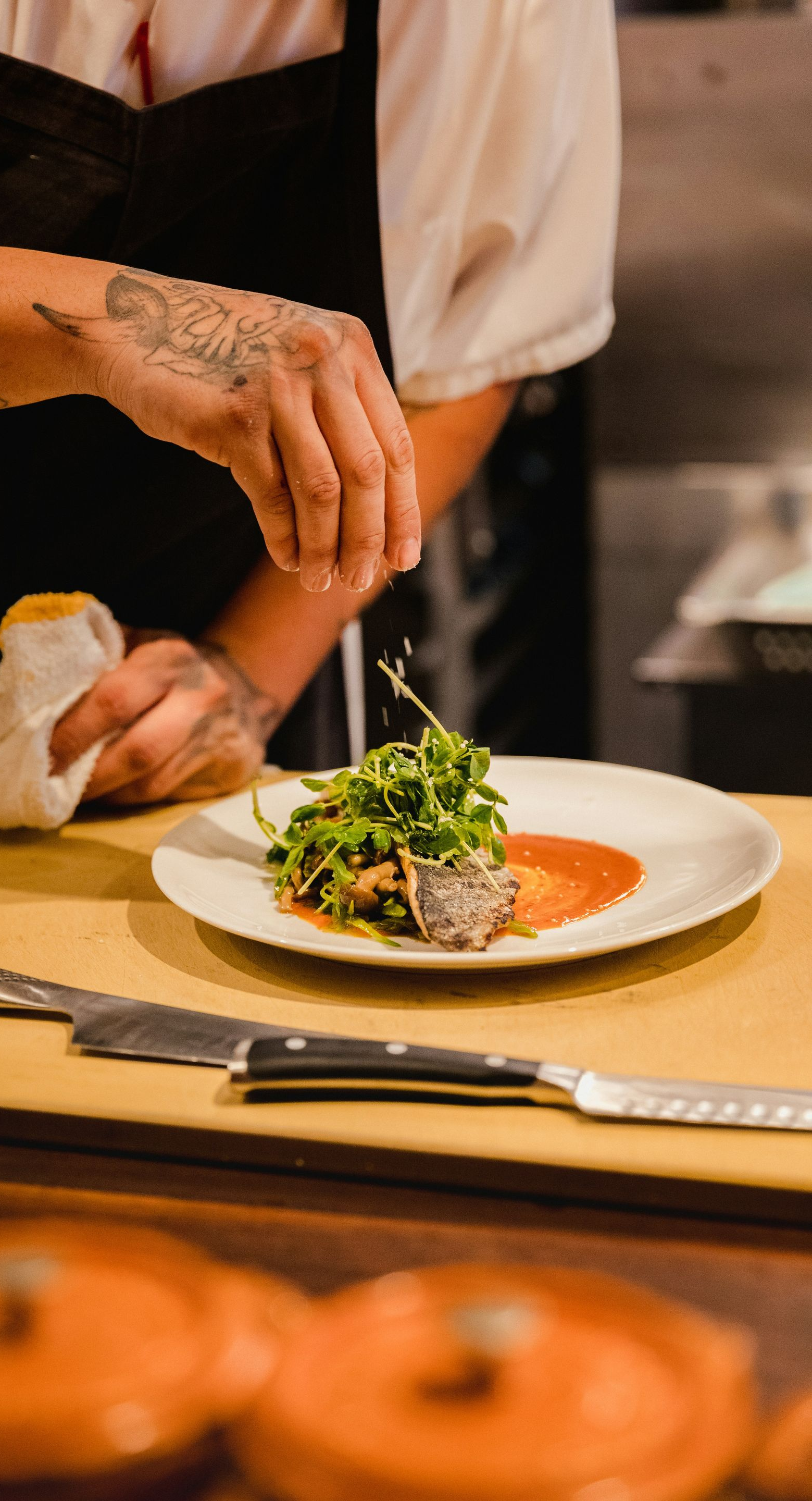Chef sprinkling seasoning onto a plated dish with arugula and sauce in a restaurant setting.