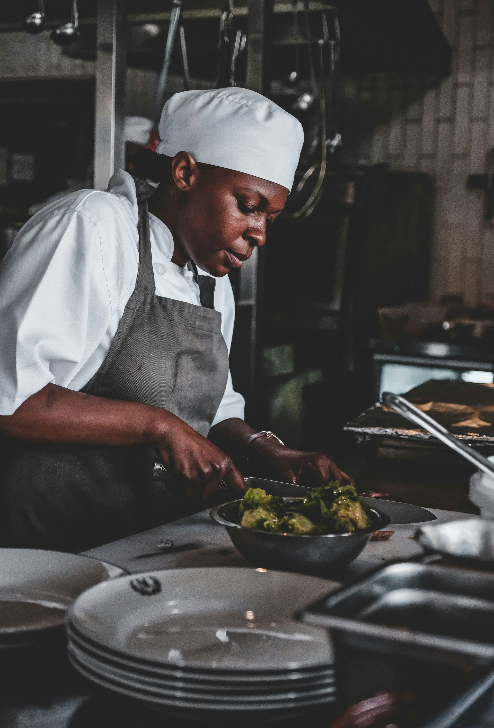 Chef in a white hat and apron cutting vegetables in a stainless steel bowl in a restaurant kitchen.