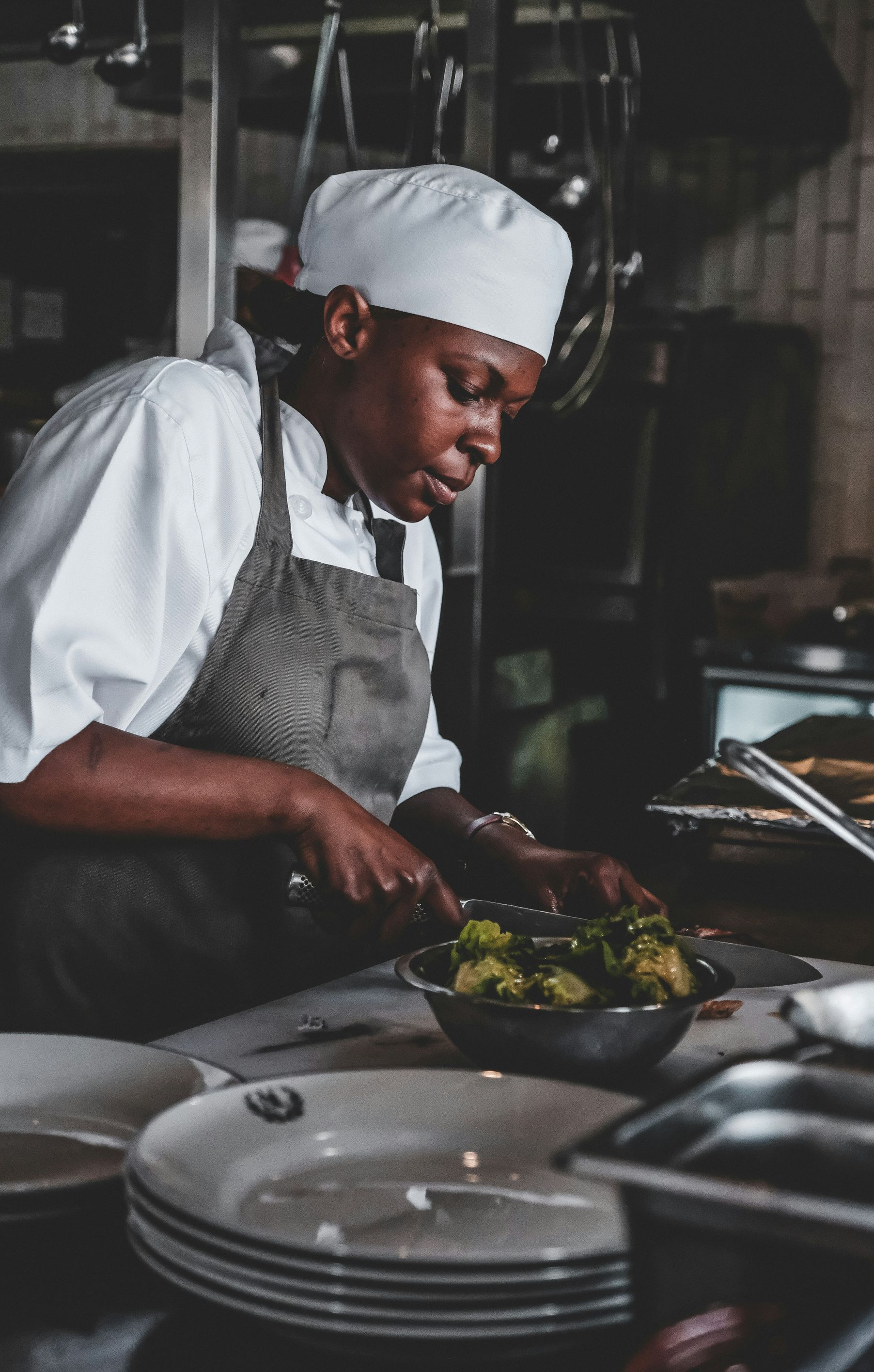Chef in a white hat and apron cutting vegetables in a stainless steel bowl in a restaurant kitchen.