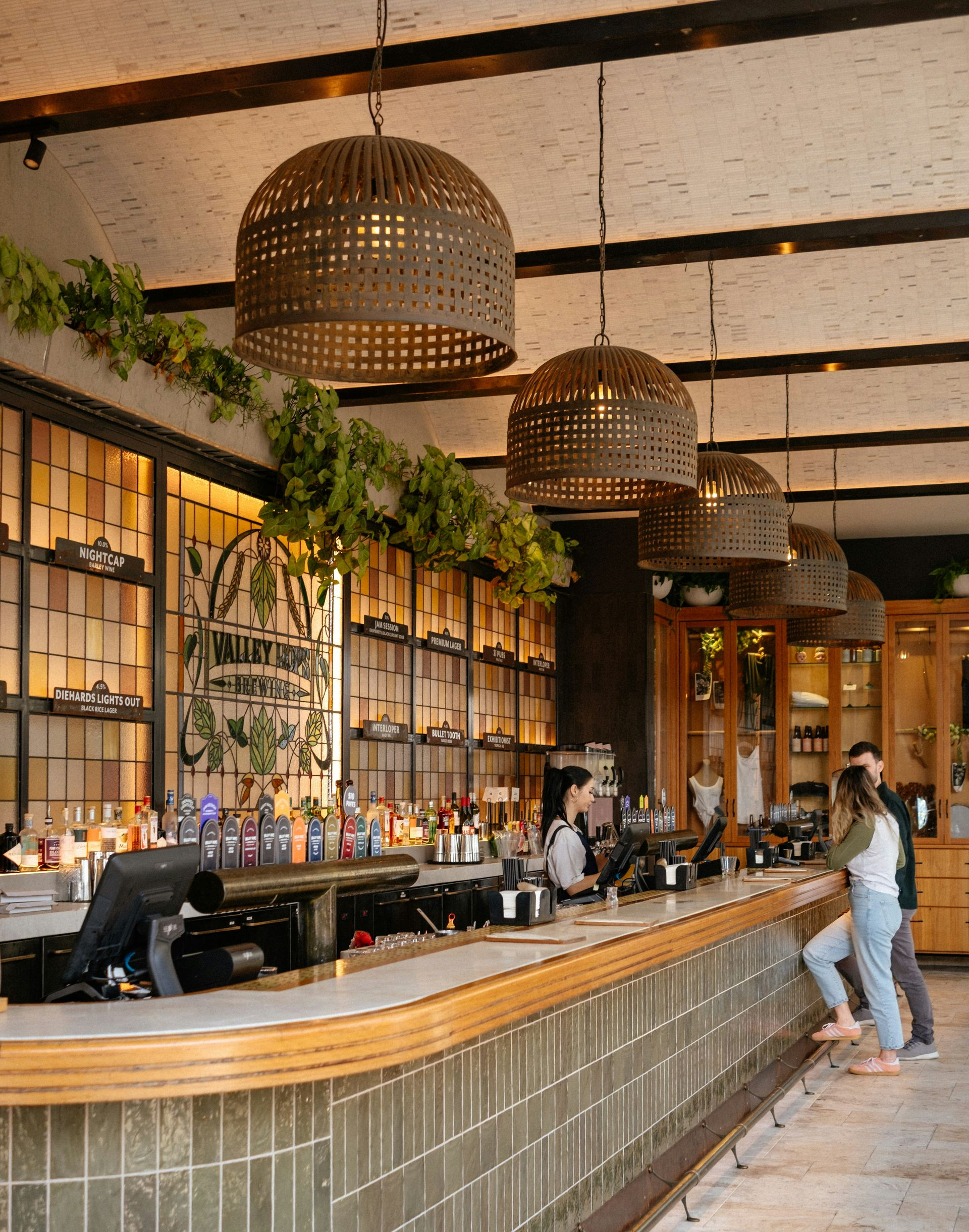 Rustic restaurant bar with wicker pendant lights, long counter, and two patrons standing at the end.