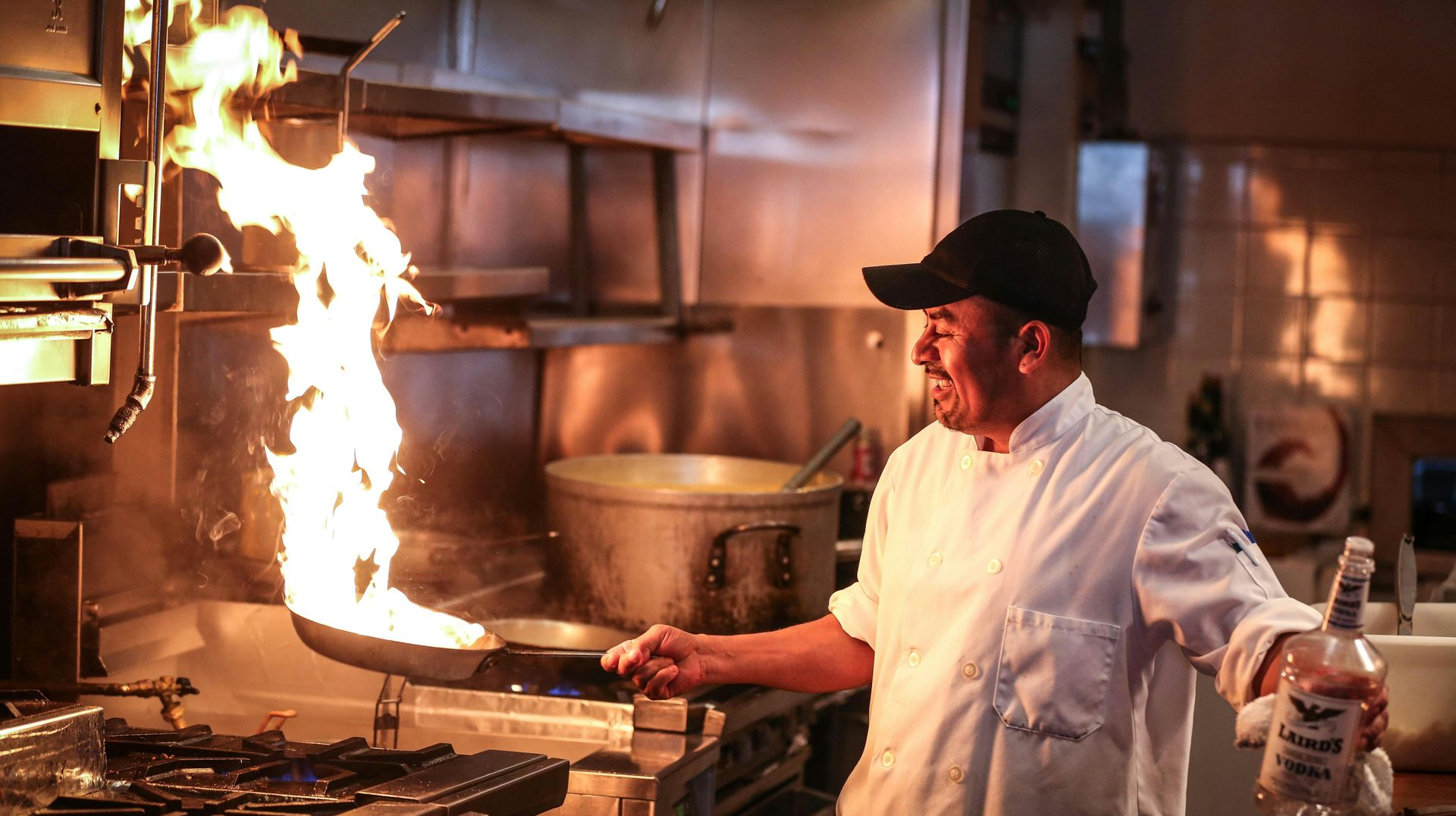Chef flambéing food in a pan, flames rising. Kitchen setting with equipment and bottle of liquor.