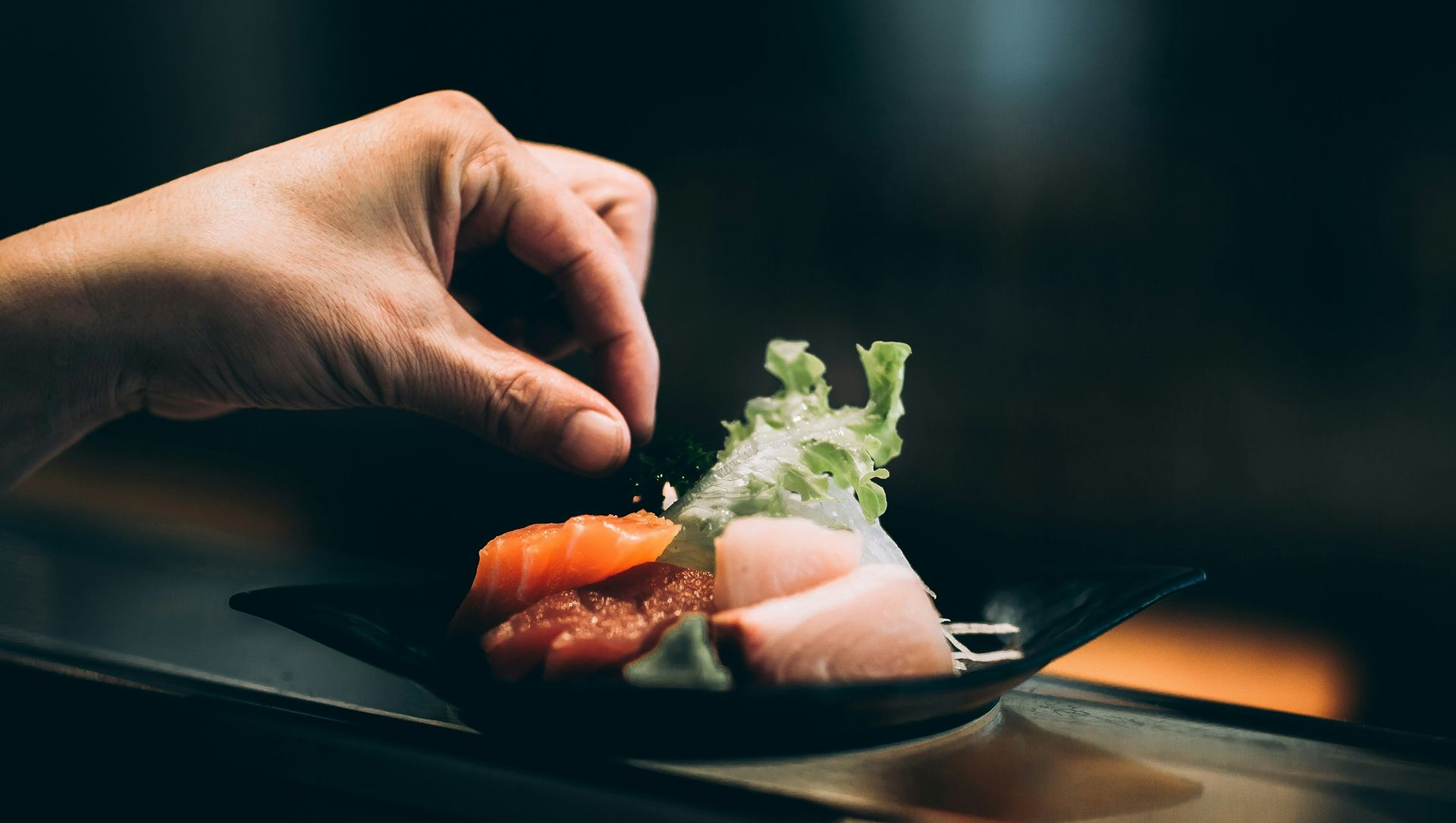 Hand garnishing sashimi on a black plate with greens.