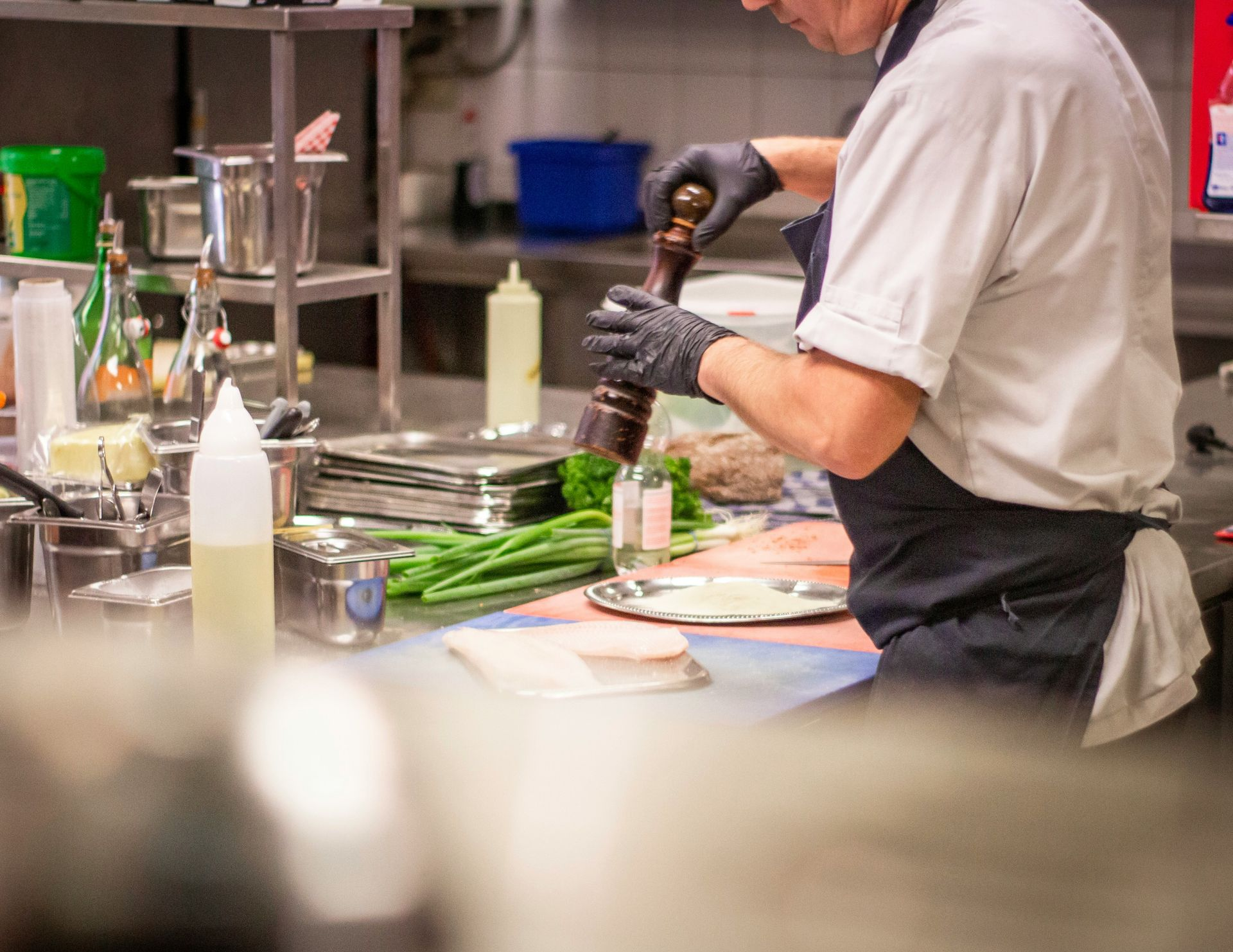 Chef in black gloves grinding pepper in a restaurant kitchen with stainless steel counters and equipment.