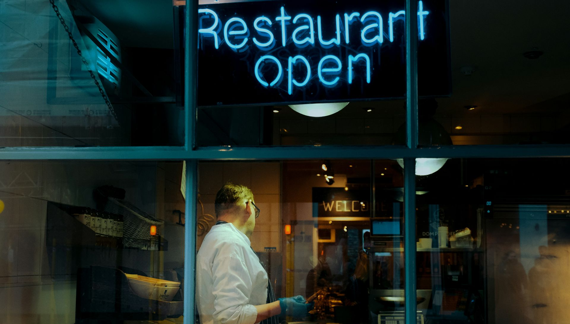 Neon “Restaurant open” sign above a person standing in a dim restaurant window at night