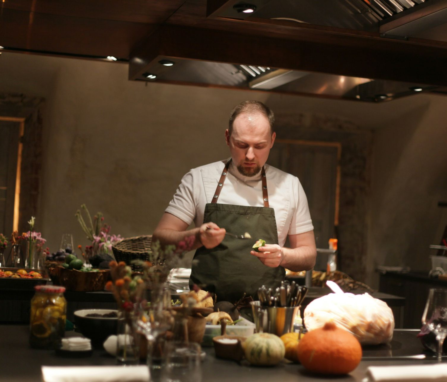 Chef plating food at a rustic kitchen counter with fall produce and floral arrangements.