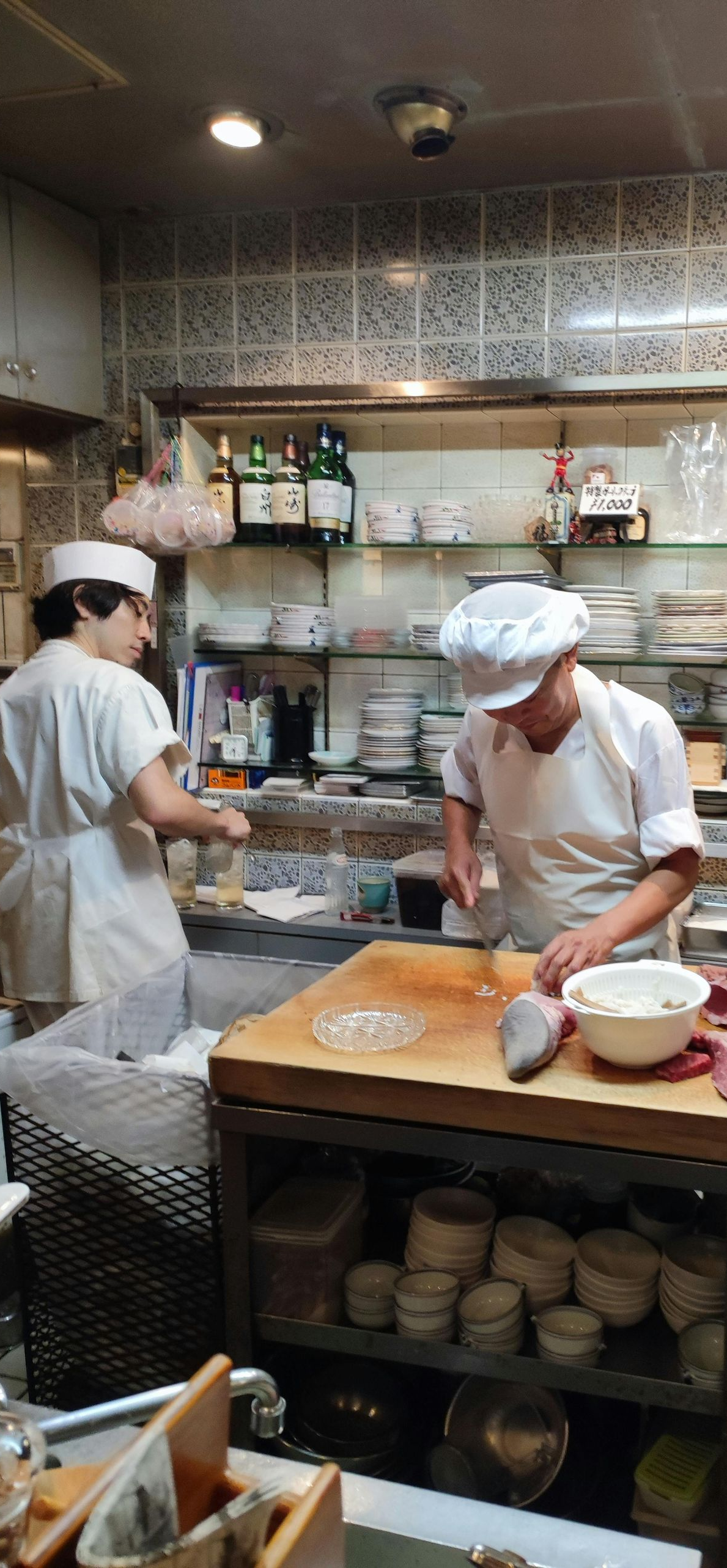 Two chefs in white uniforms and hats work behind a wooden counter in a kitchen, preparing food with a knife.