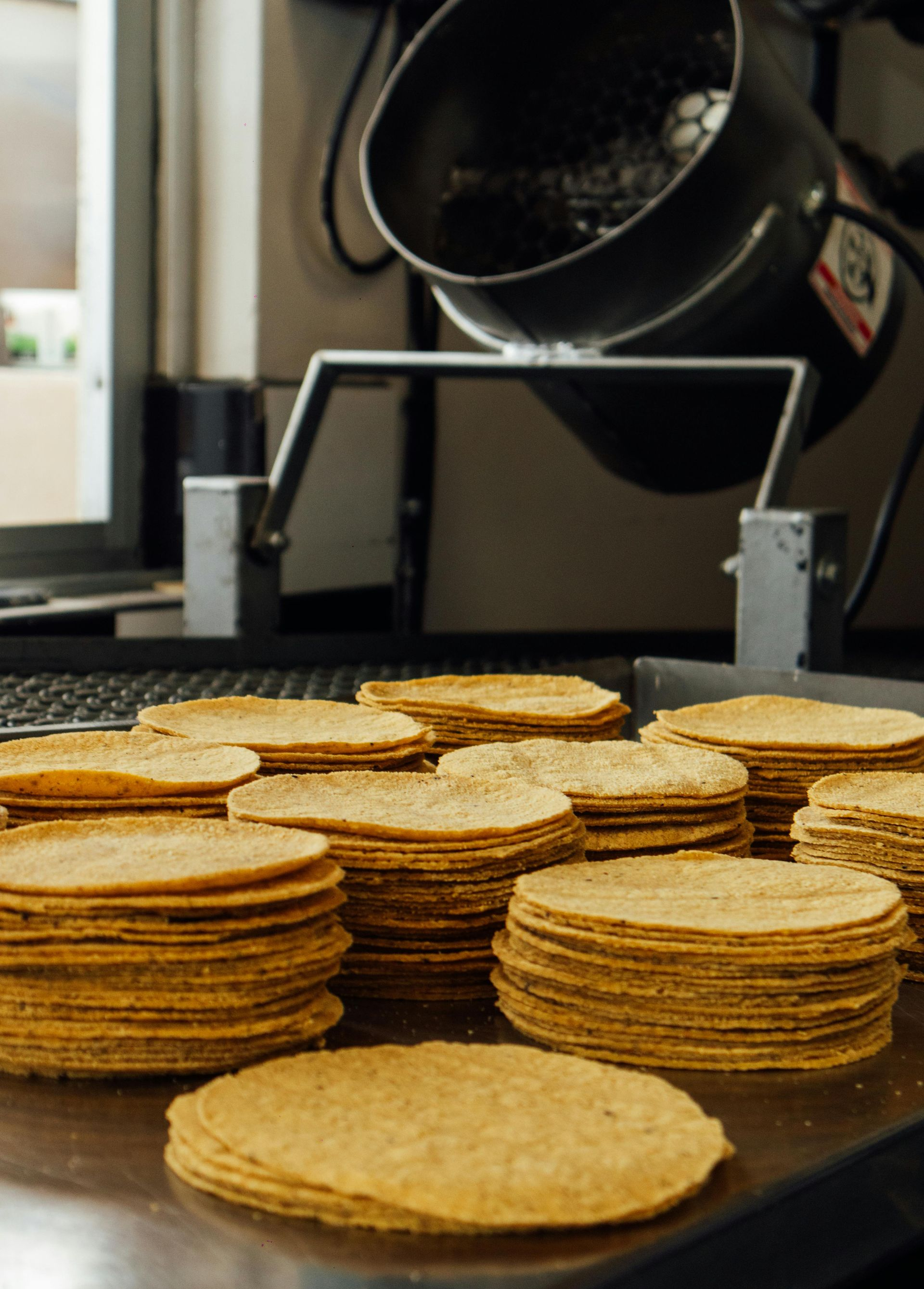 Stacks of golden-brown tortillas on a metal surface, with a tortilla-making machine in the background.