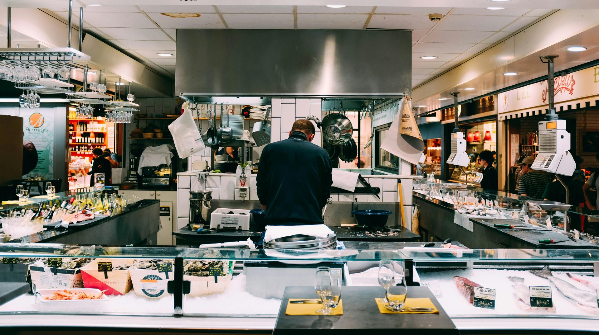 A person stands at a counter in a food market, with shelves of items and cooking area in the background.