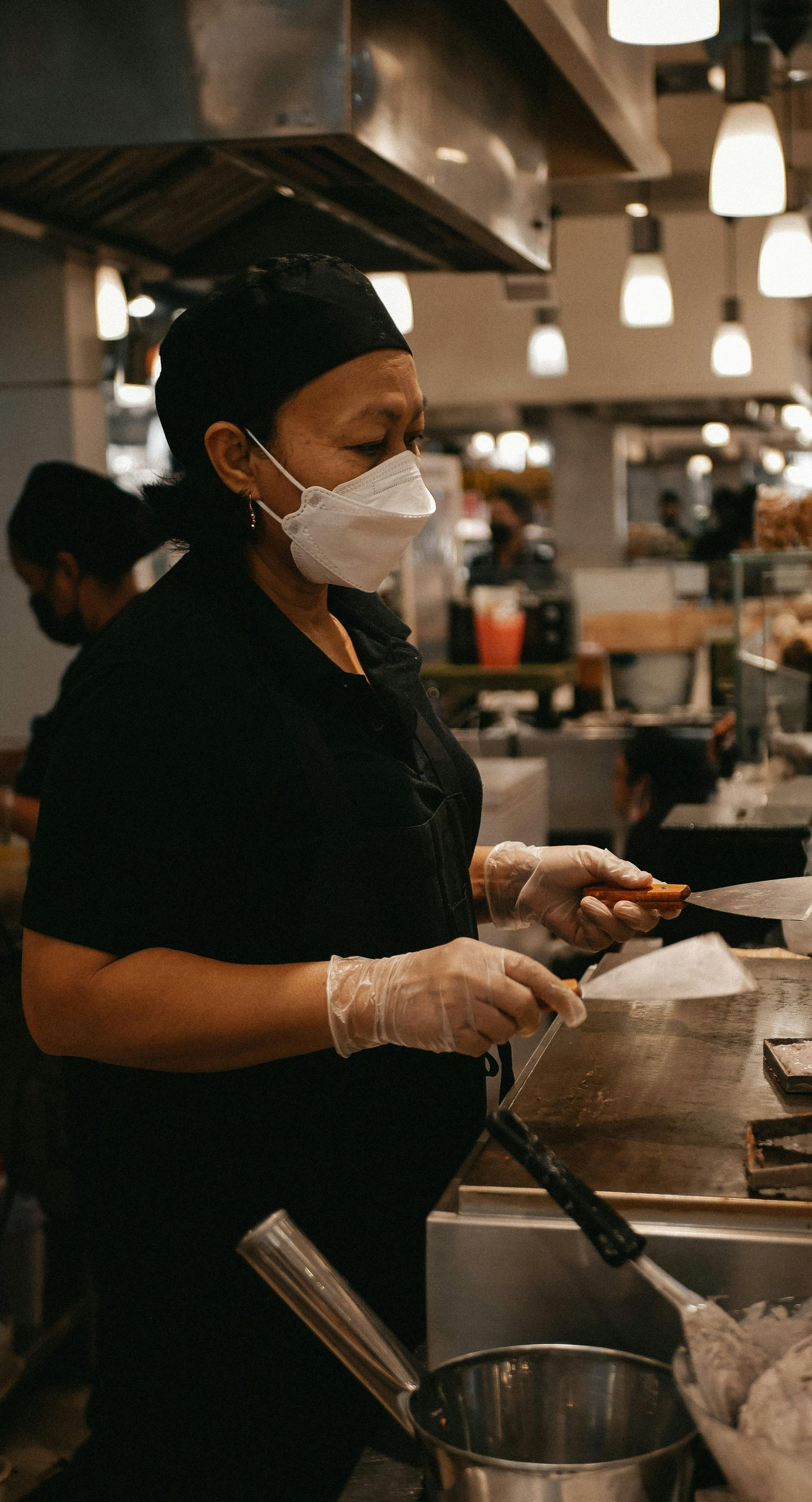 Cook in a commercial kitchen wearing a mask and gloves, using a spatula over a pot.