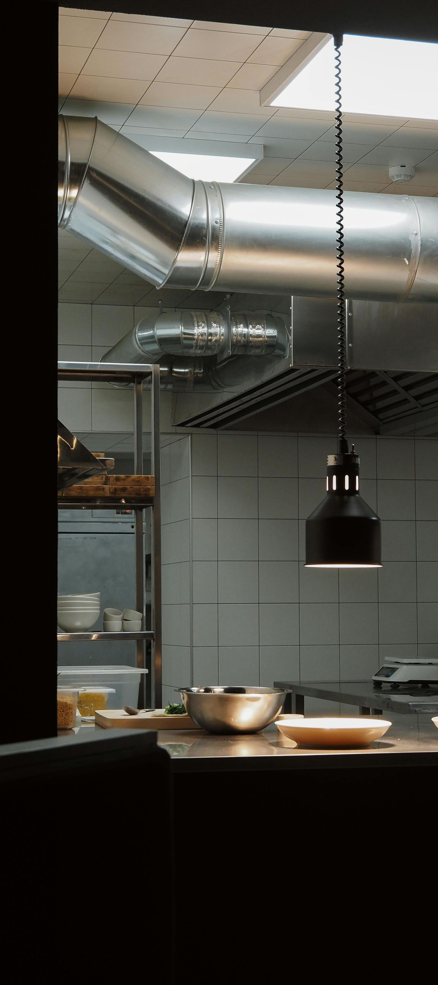 A professional kitchen interior featuring a metal ventilation duct, a hanging pendant light, and stainless steel counters.