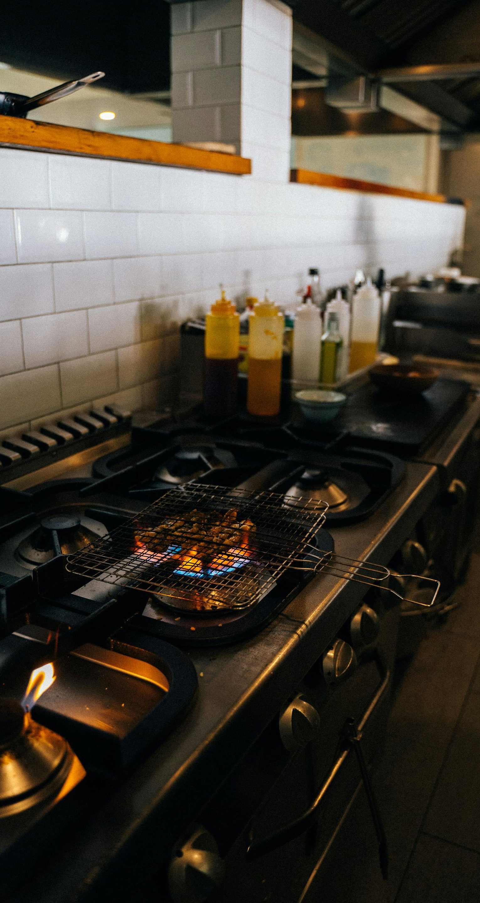A professional kitchen stove with gas burners lit, next to various squeeze bottles on a white tiled counter.