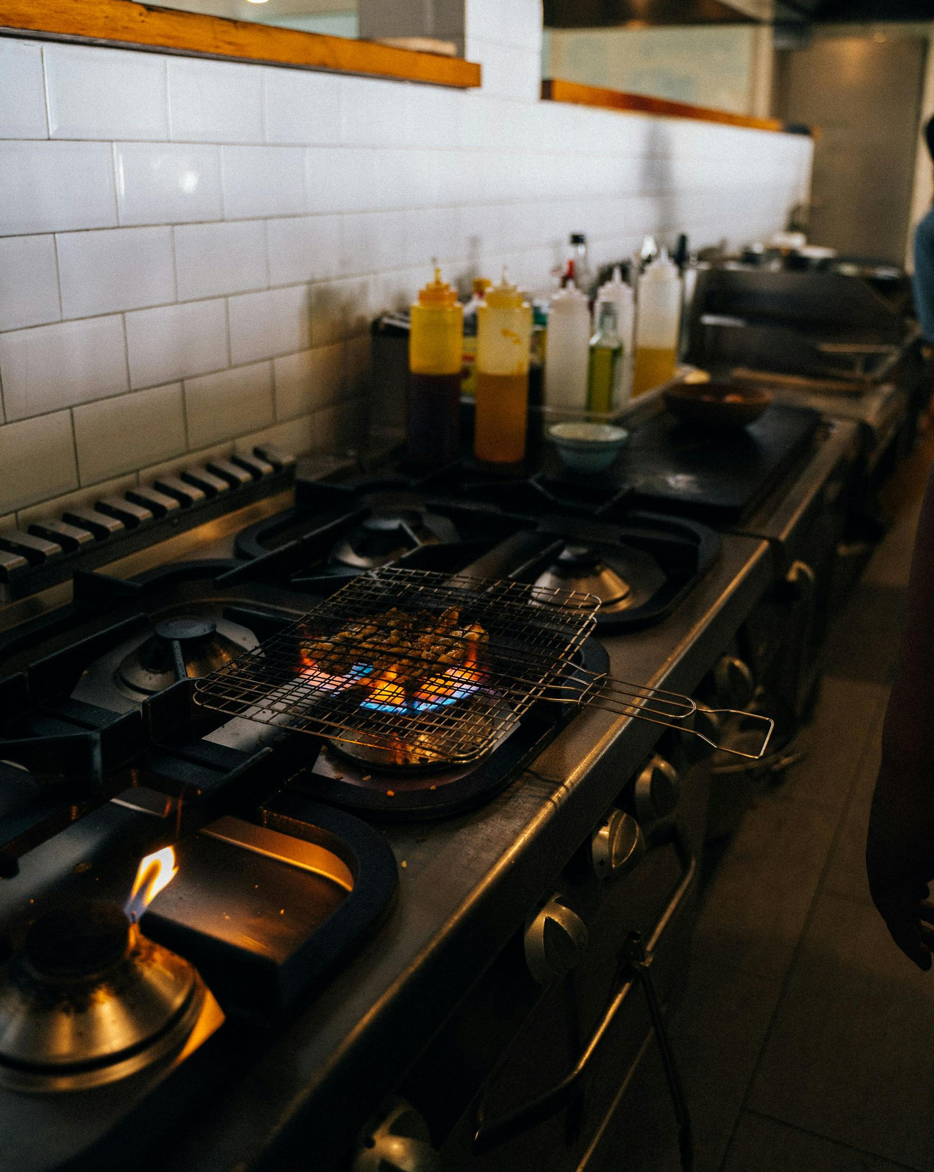 Commercial kitchen stove with flames. Bottles and pans sit on the stove; white-tiled wall in background.