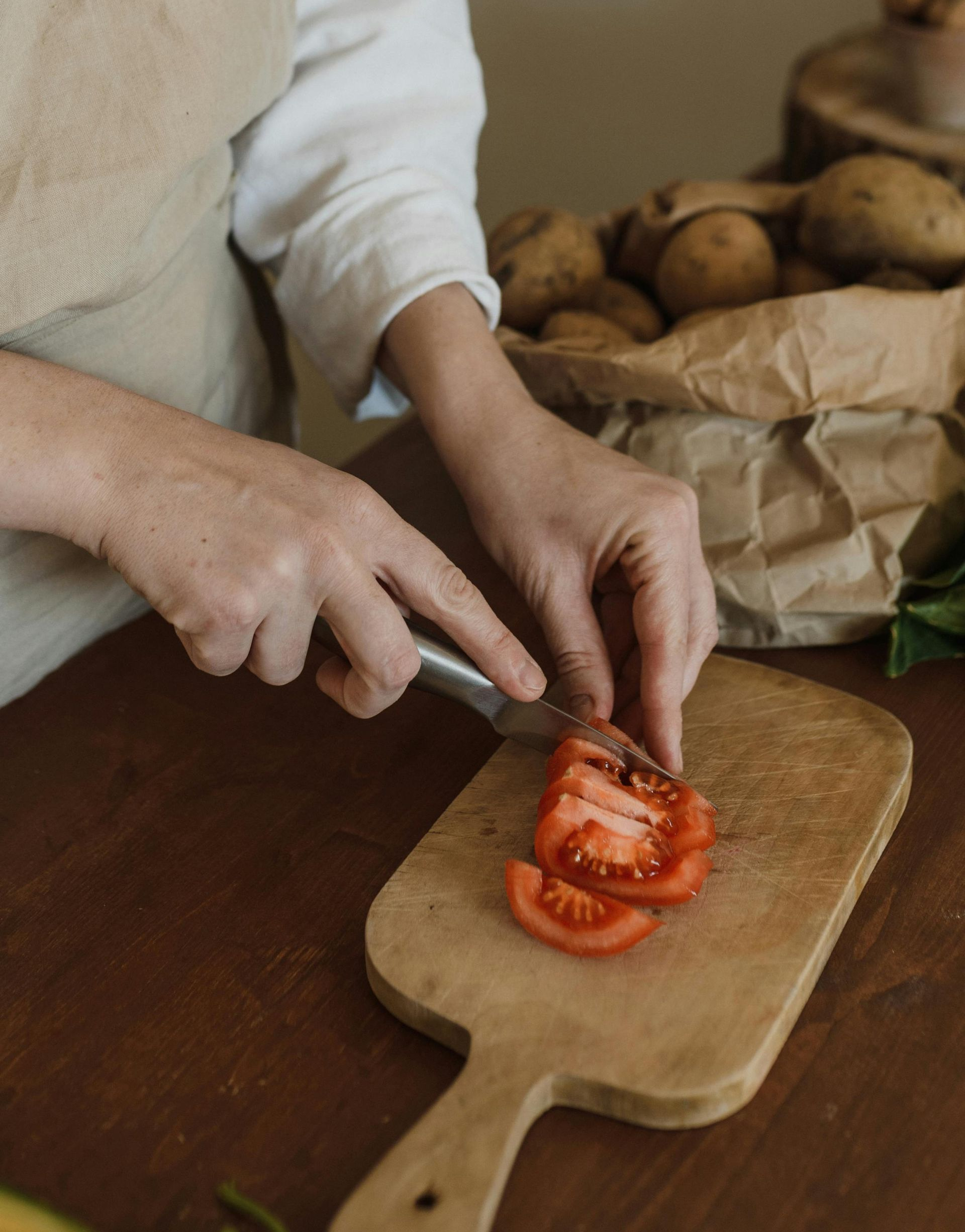 A person in a beige apron slices a tomato on a wooden cutting board next to a paper bag filled with potatoes.