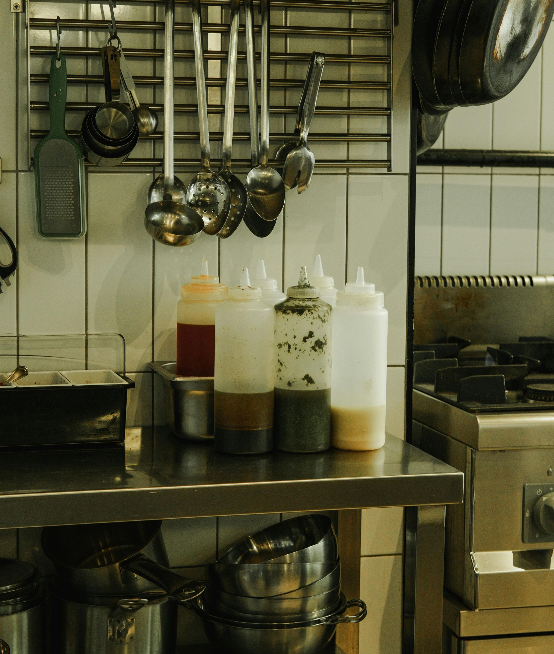 A commercial kitchen prep area with metal utensils hanging on a rack above four condiment squeeze bottles on a counter.