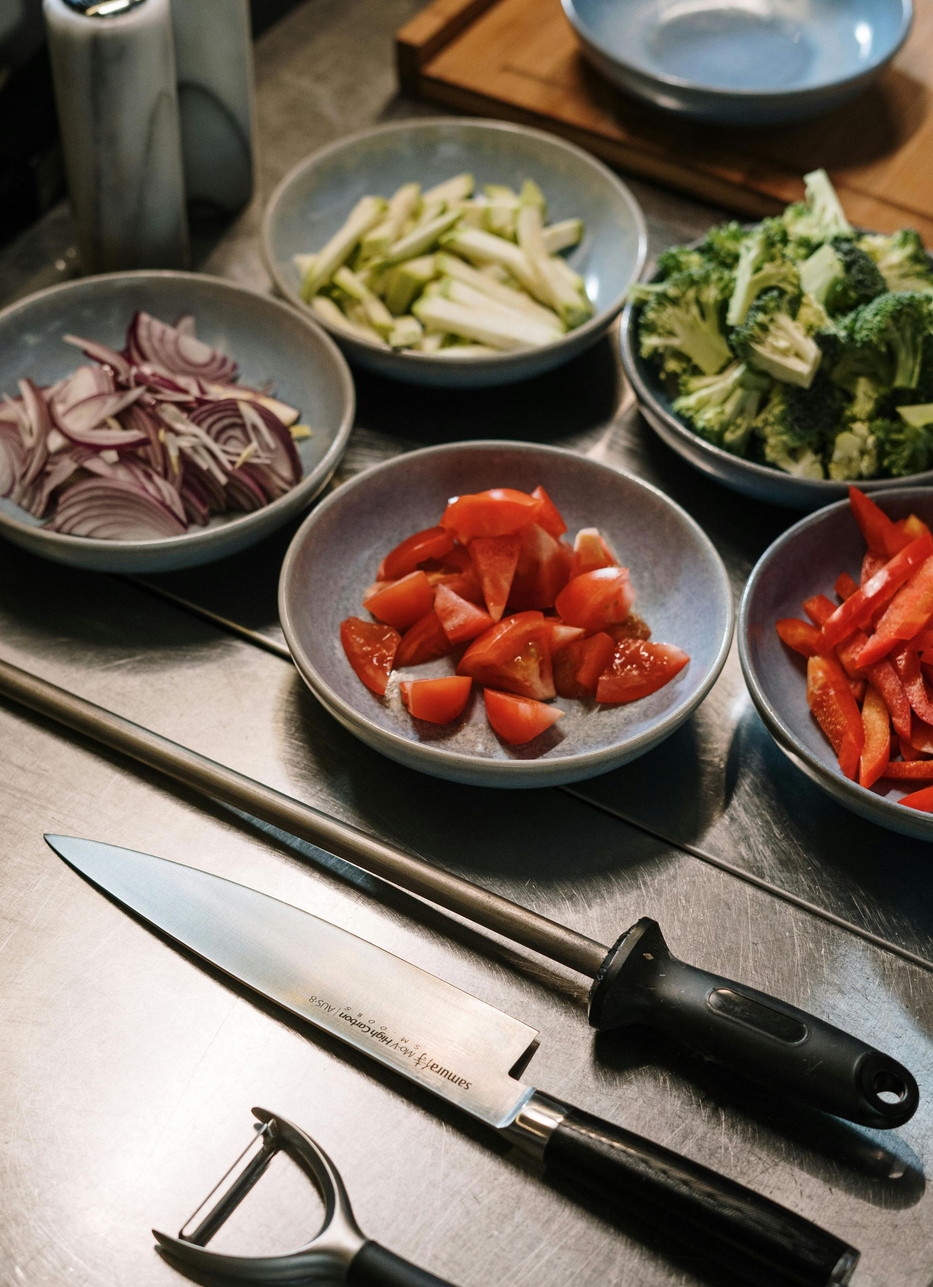 A kitchen counter displays various bowls of chopped vegetables, a chef's knife, a sharpening rod, and a vegetable peeler.