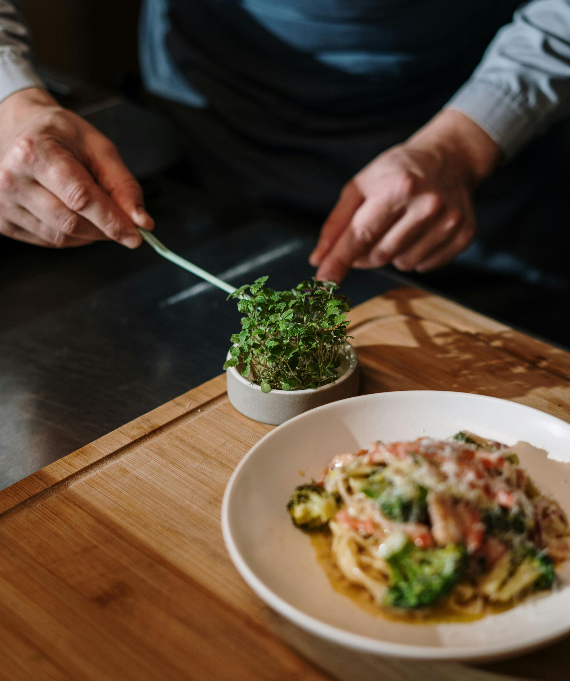A chef uses tweezers to place fresh microgreens on a white plate of pasta topped with broccoli and melted cheese.