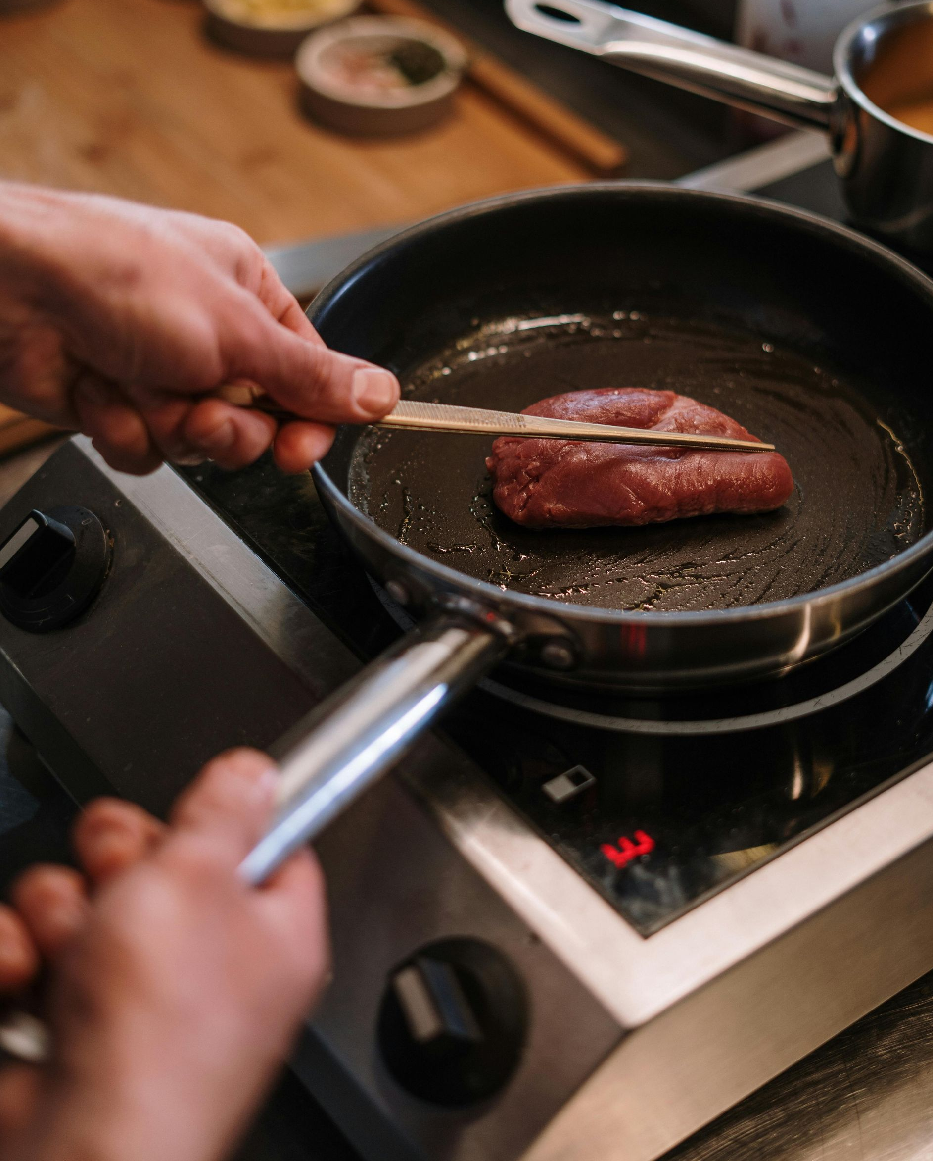 A hand holds a stick to turn a steak cooking in a pan on a stovetop.